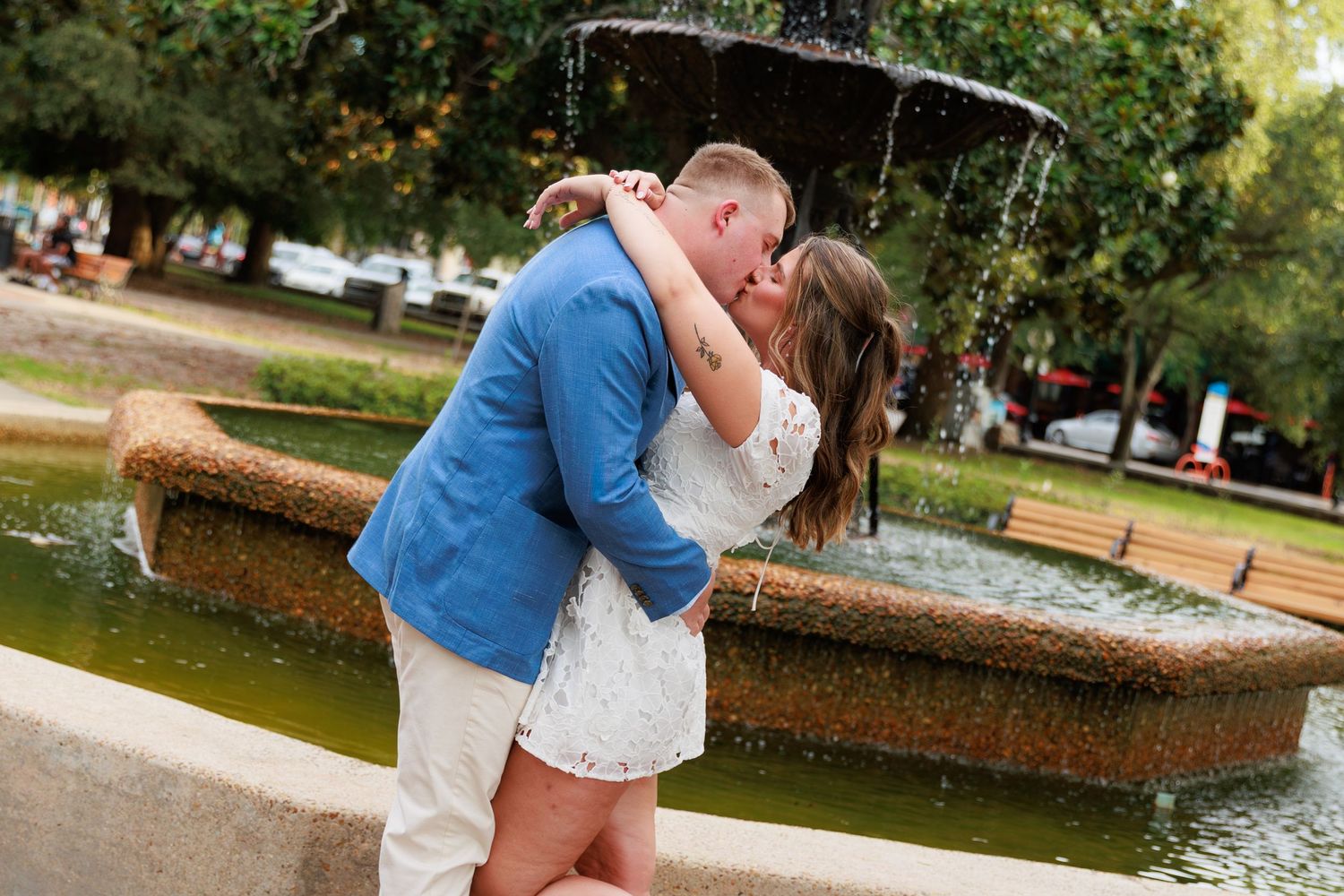 Couple sharing a romantic kiss in front of a garden fountain in a lush green park setting.