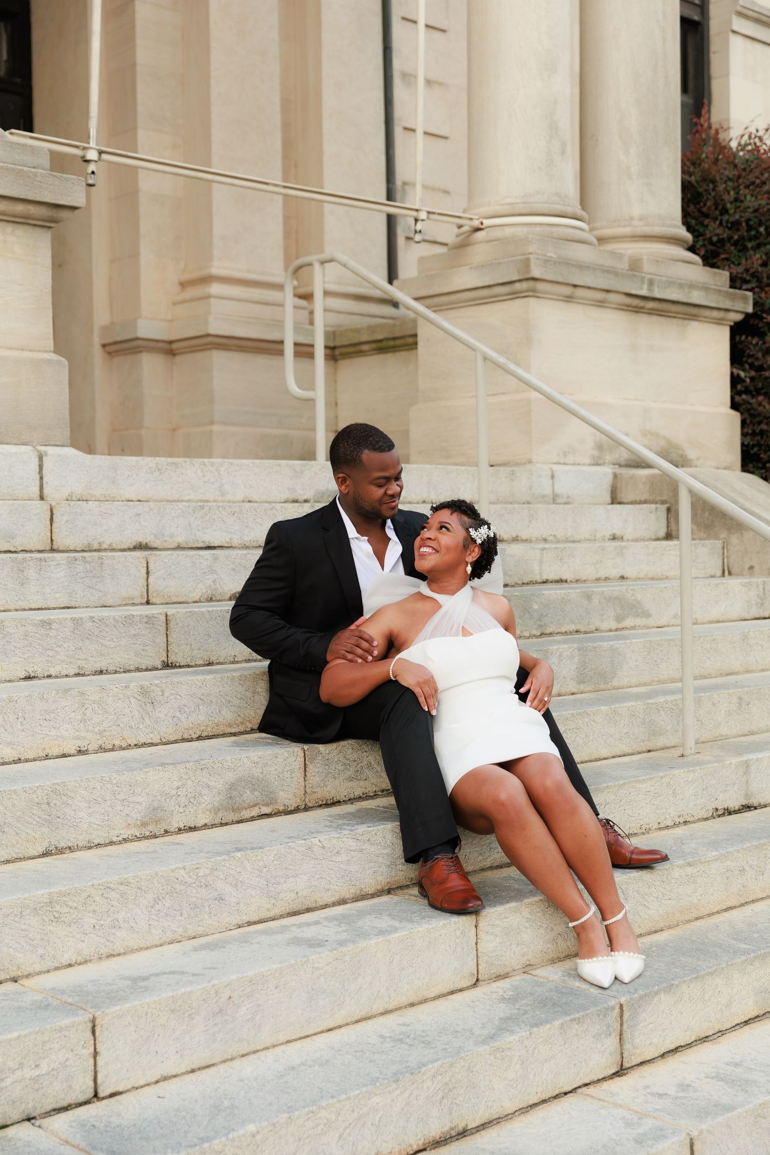 A couple in wedding attire sits on stone steps, the bride in a white mini dress smiling up at the groom in a black suit.