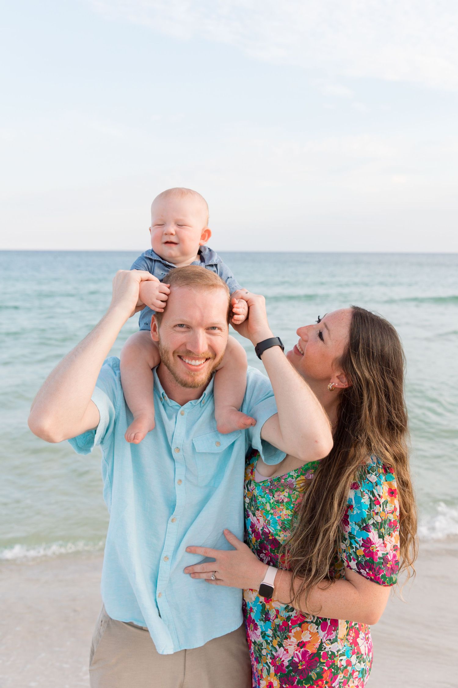 Happy family with baby on dad's shoulders smiling at the beach with turquoise ocean waves in the background.