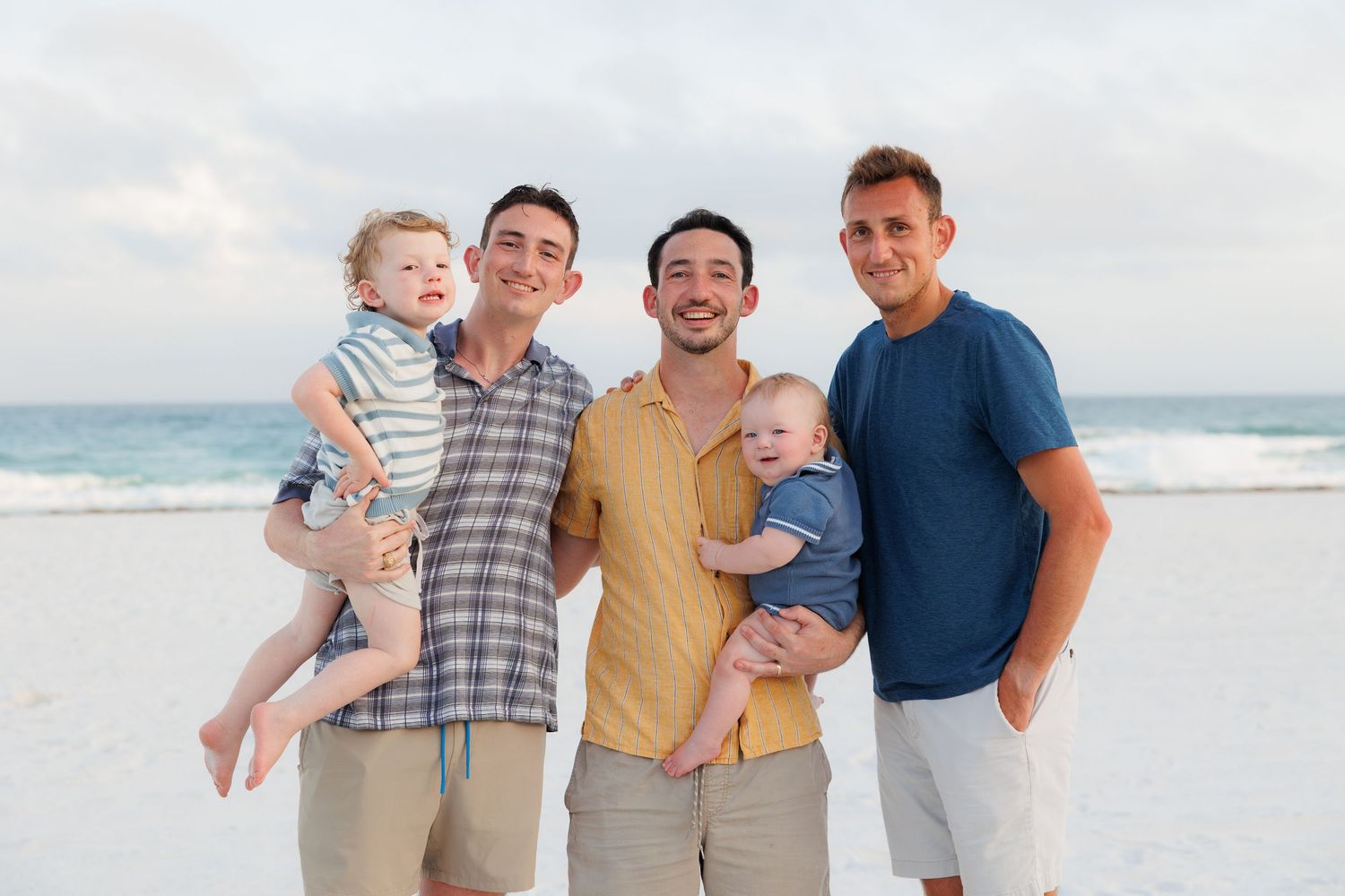 Three men holding two babies pose together smiling on a sandy beach with ocean waves in the background.