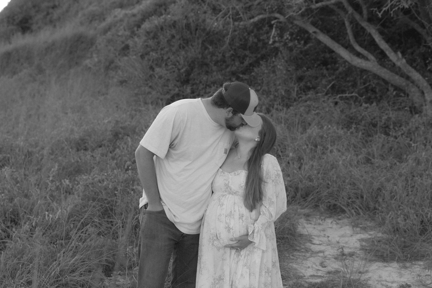 Black and white photo of a pregnant couple kissing outdoors on a nature path surrounded by lush greenery.