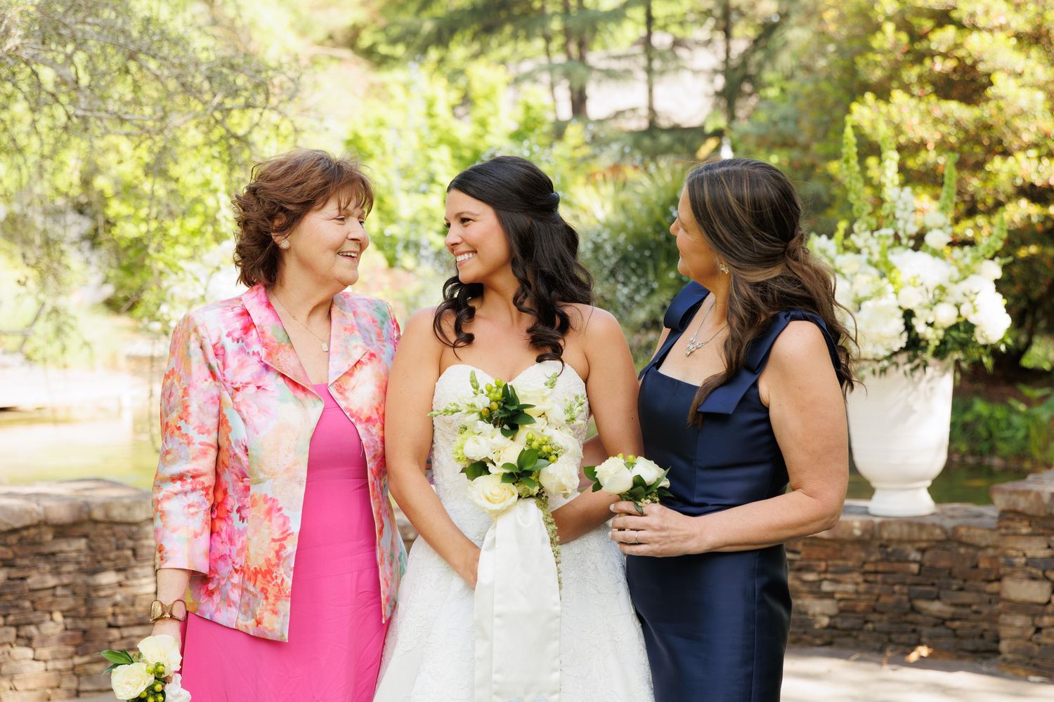 Bride in white gown holds bouquet flanked by mother in pink floral jacket and bridesmaid in navy dress outdoors.