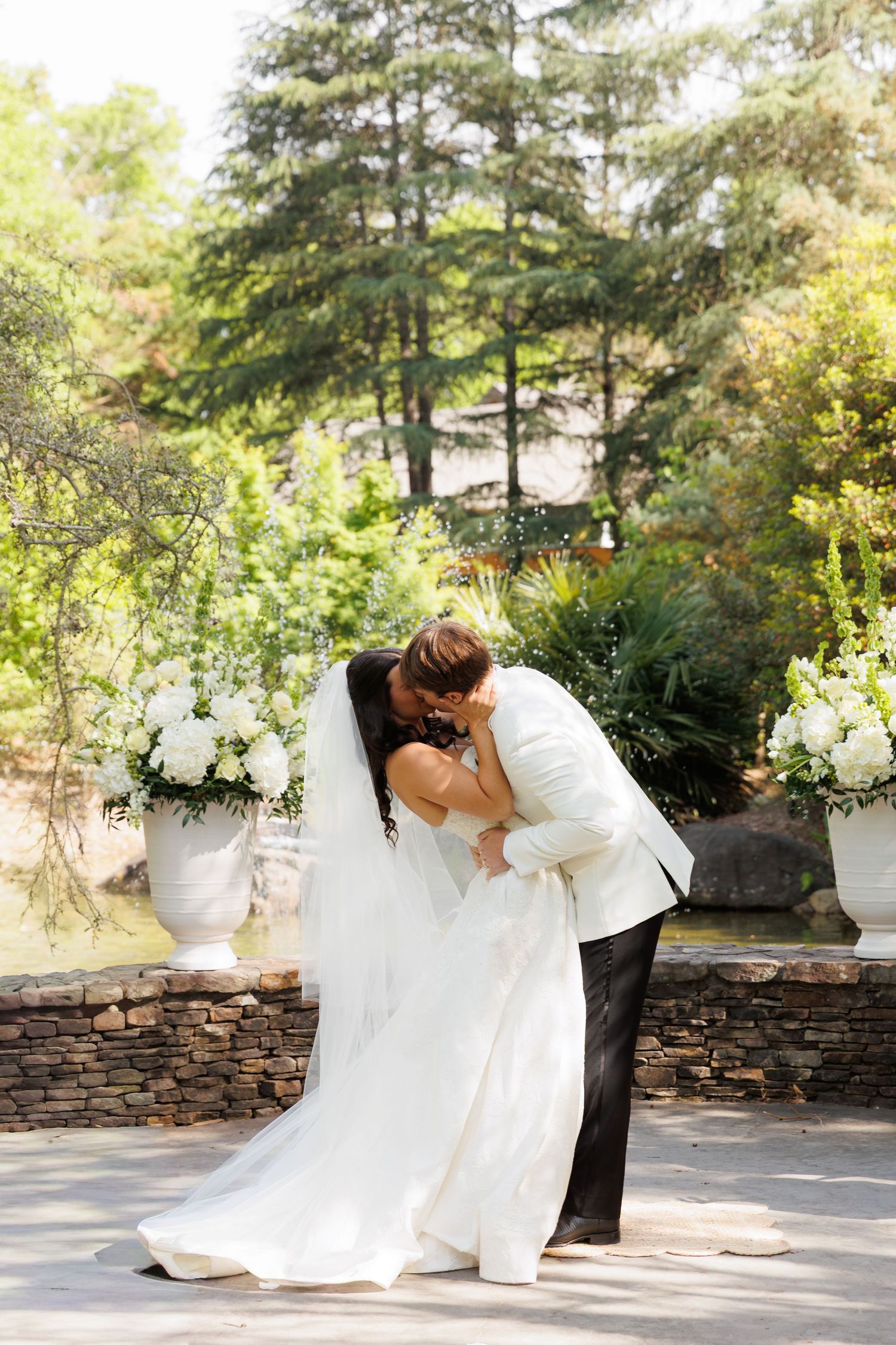 Bride and groom share romantic dip kiss surrounded by white floral arrangements in garden ceremony.