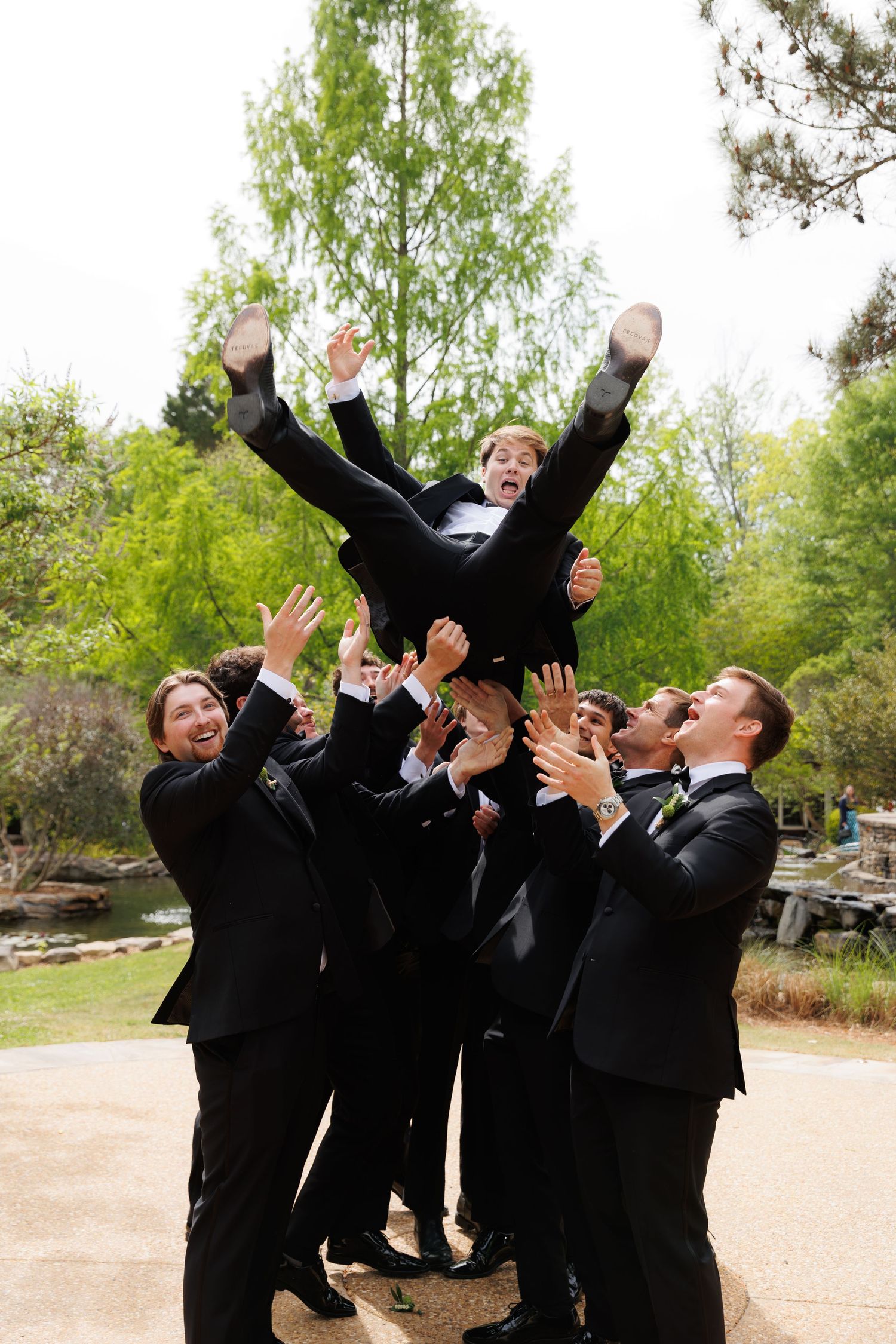 Groomsmen in black tuxedos toss groom into the air outdoors at wedding celebration.