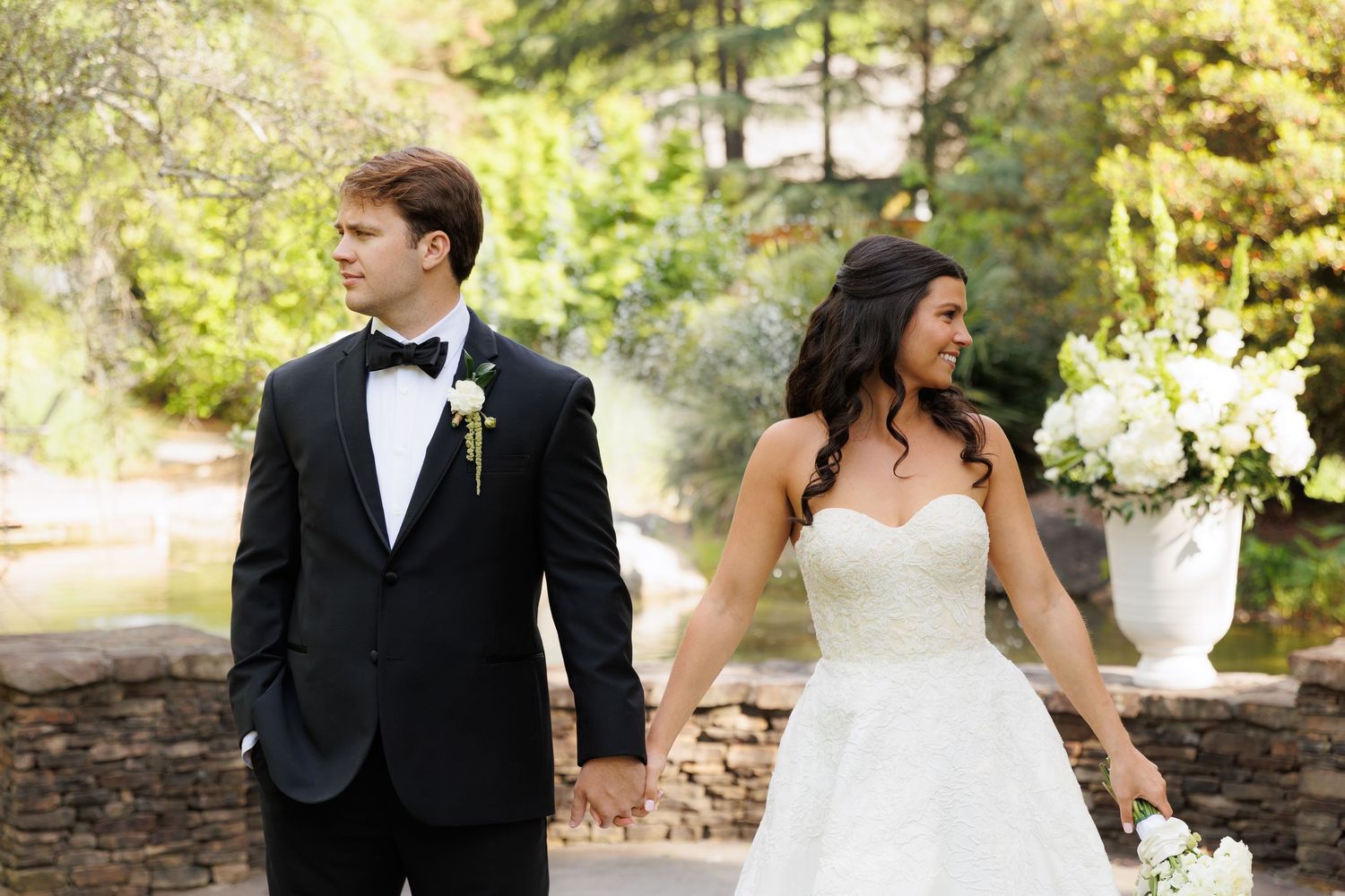 Bride and groom hold hands in elegant black tuxedo and white strapless gown outdoors.