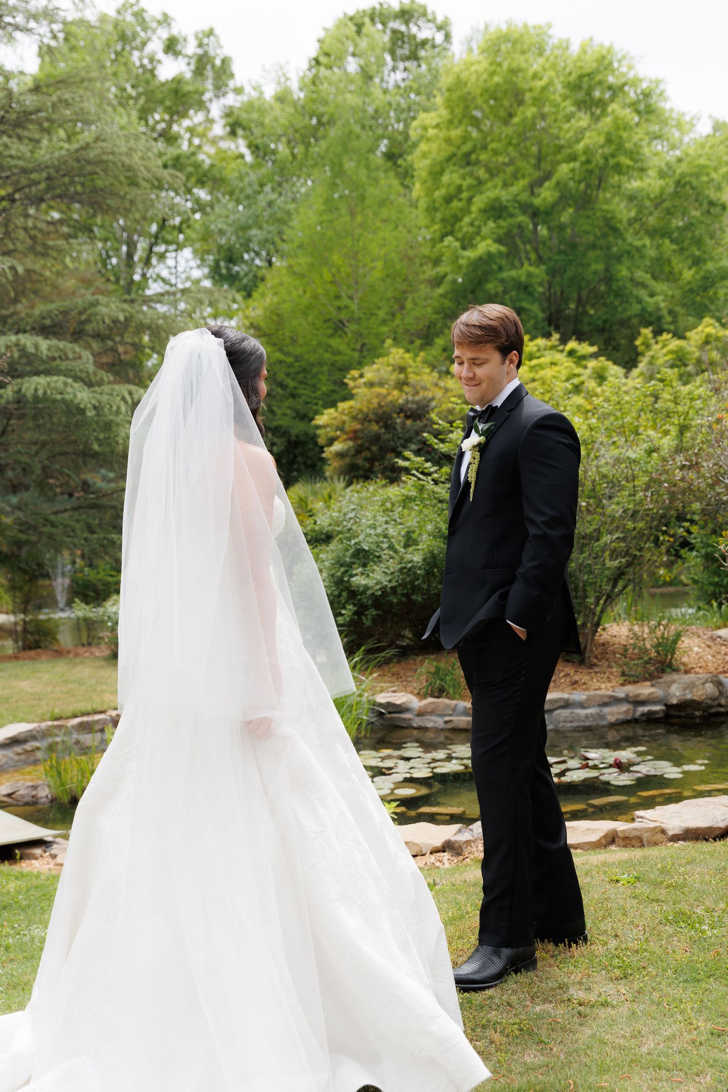 Bride in white gown and veil faces groom in black tuxedo during outdoor wedding ceremony in lush garden.