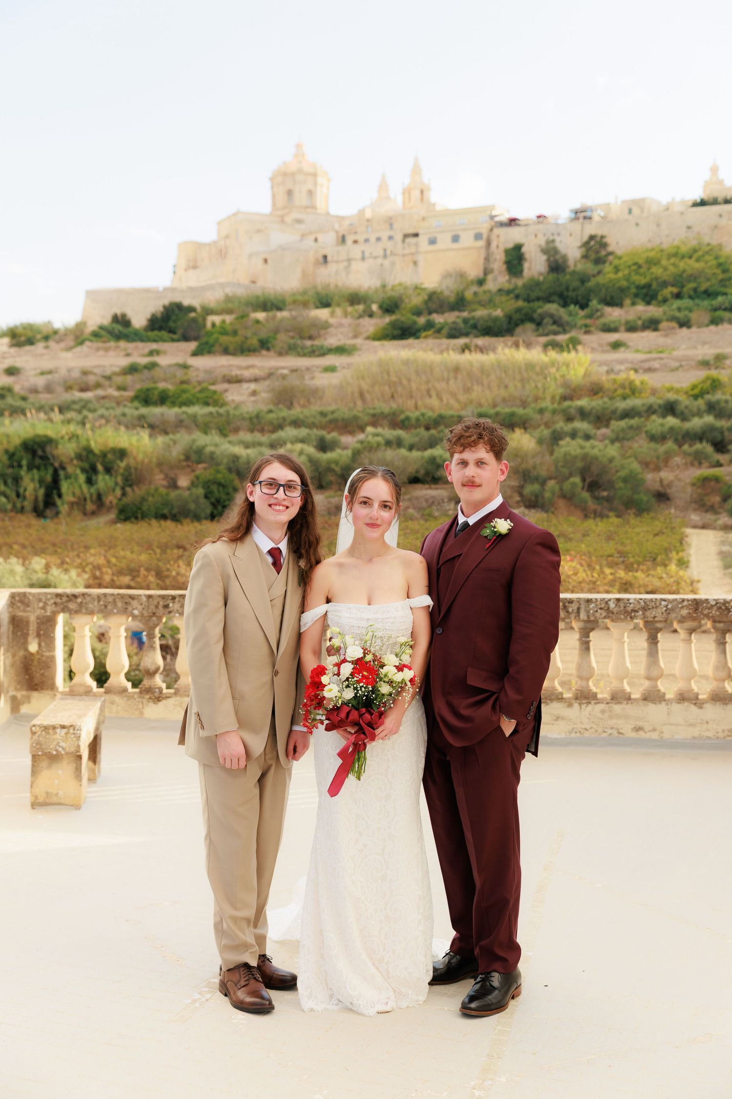 A wedding party poses on a stone terrace with ancient fortress walls and greenery in the background.
