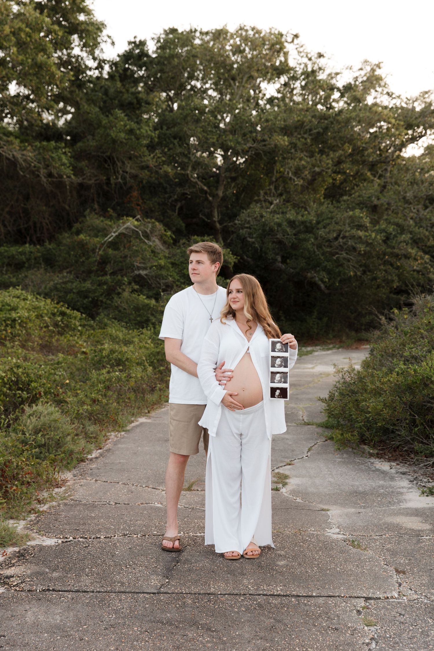 A couple in white clothing embraces while walking along a gravel path surrounded by greenery.