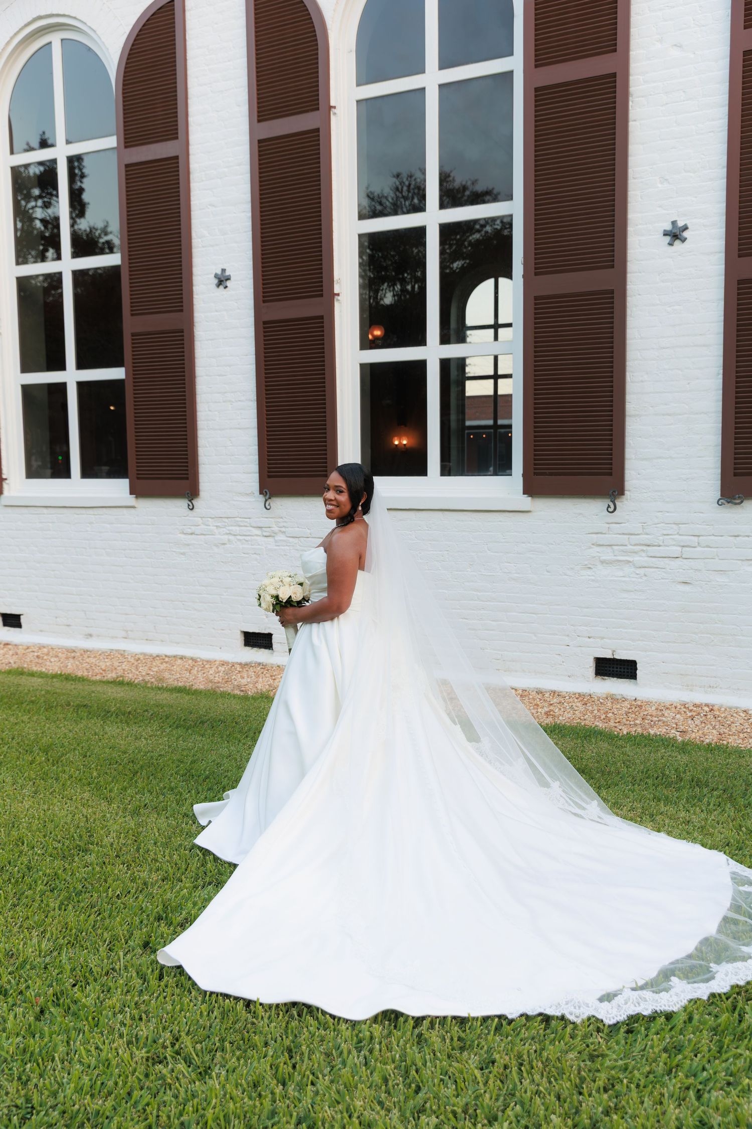 Flowing white wedding dress train spreads beautifully across green lawn near white church building.