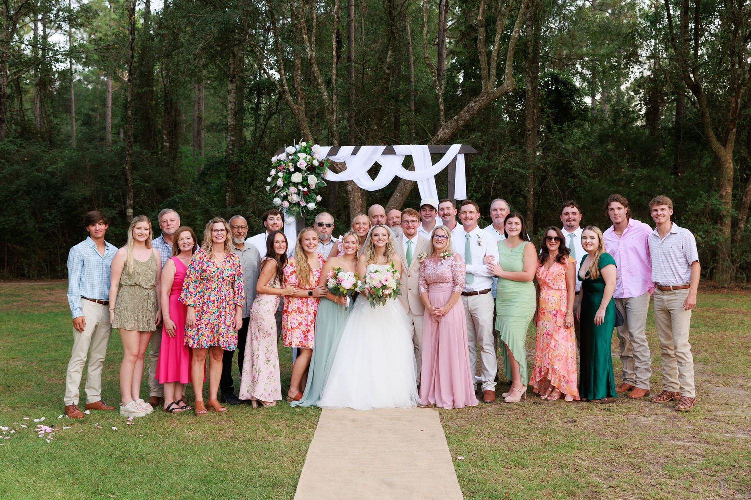 Wedding party in pink dresses and khaki suits poses together outdoors in a wooded setting.