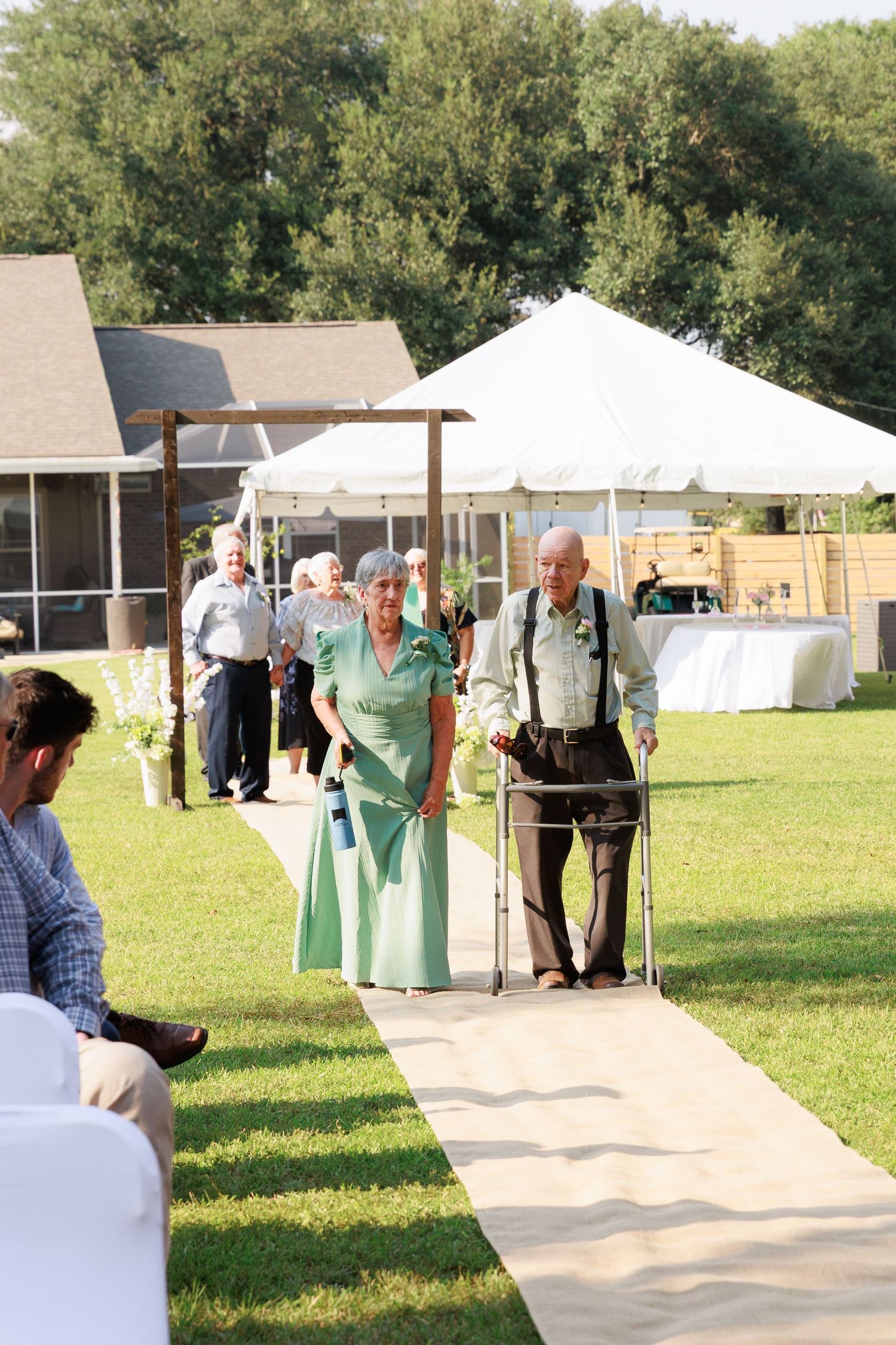 Wedding guests walk down a concrete path at an outdoor venue with white tents and green grass in the background.