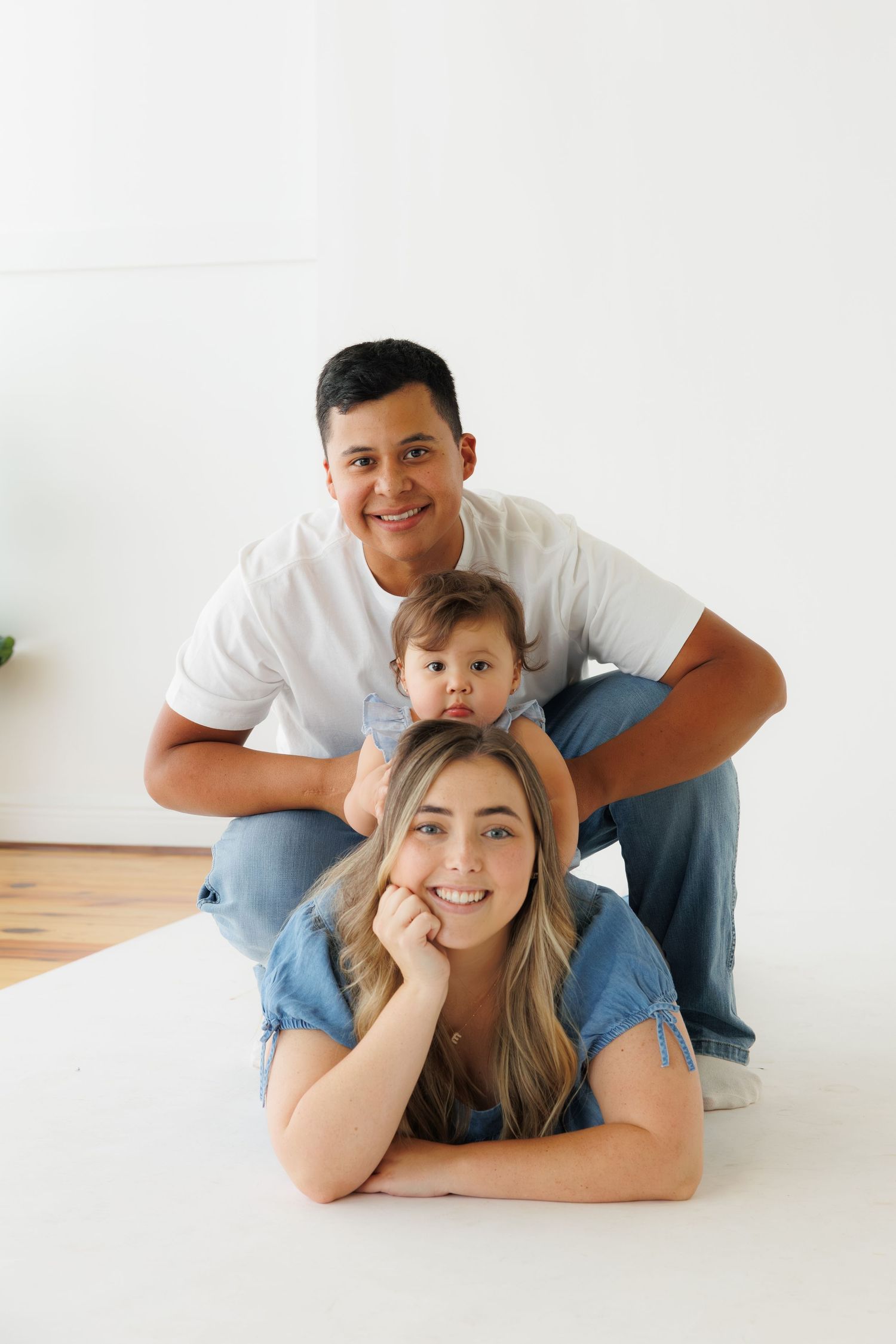 Family of three posing on floor, parents in denim and white, toddler between them, smiling at camera.