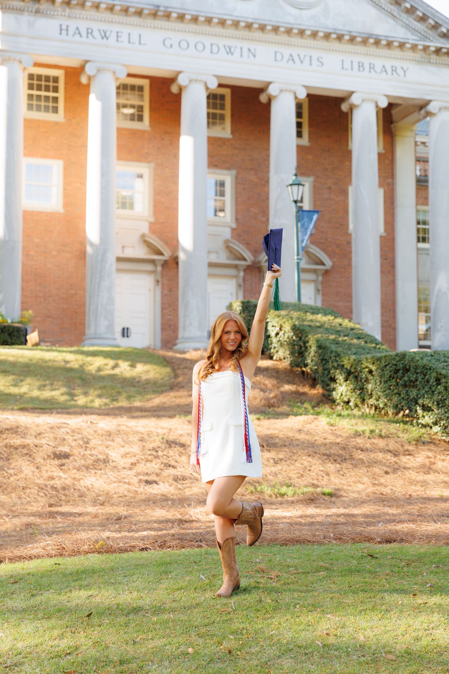 Smiling graduate in white dress and cowboy boots raises cap in front of Harwell Goodwin Davis Library.