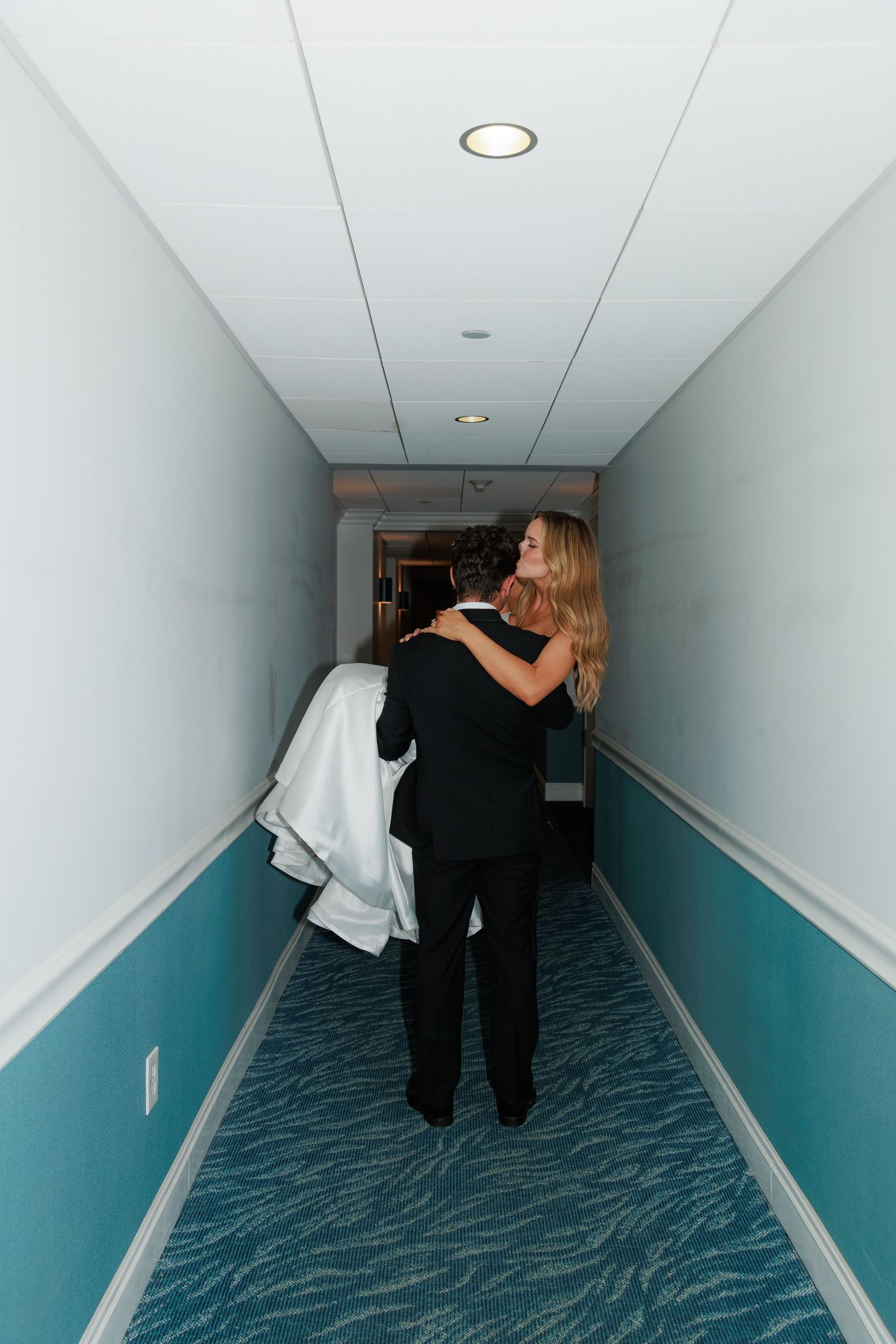 A tender embrace between two people in a blue-walled hotel hallway with patterned carpet.