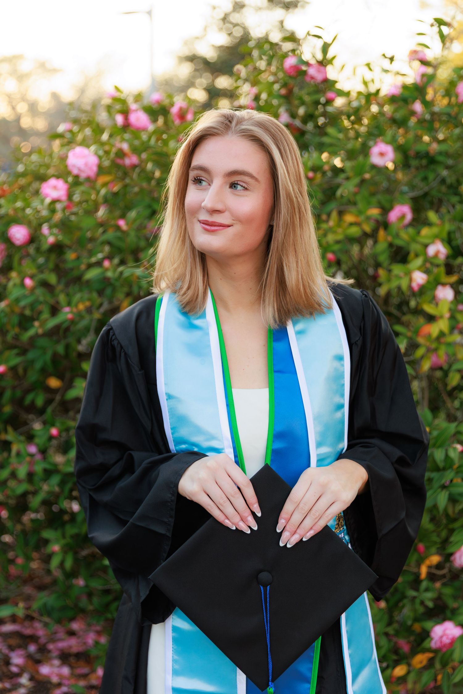 Graduate in academic regalia poses with light blue stole against a backdrop of pink roses.