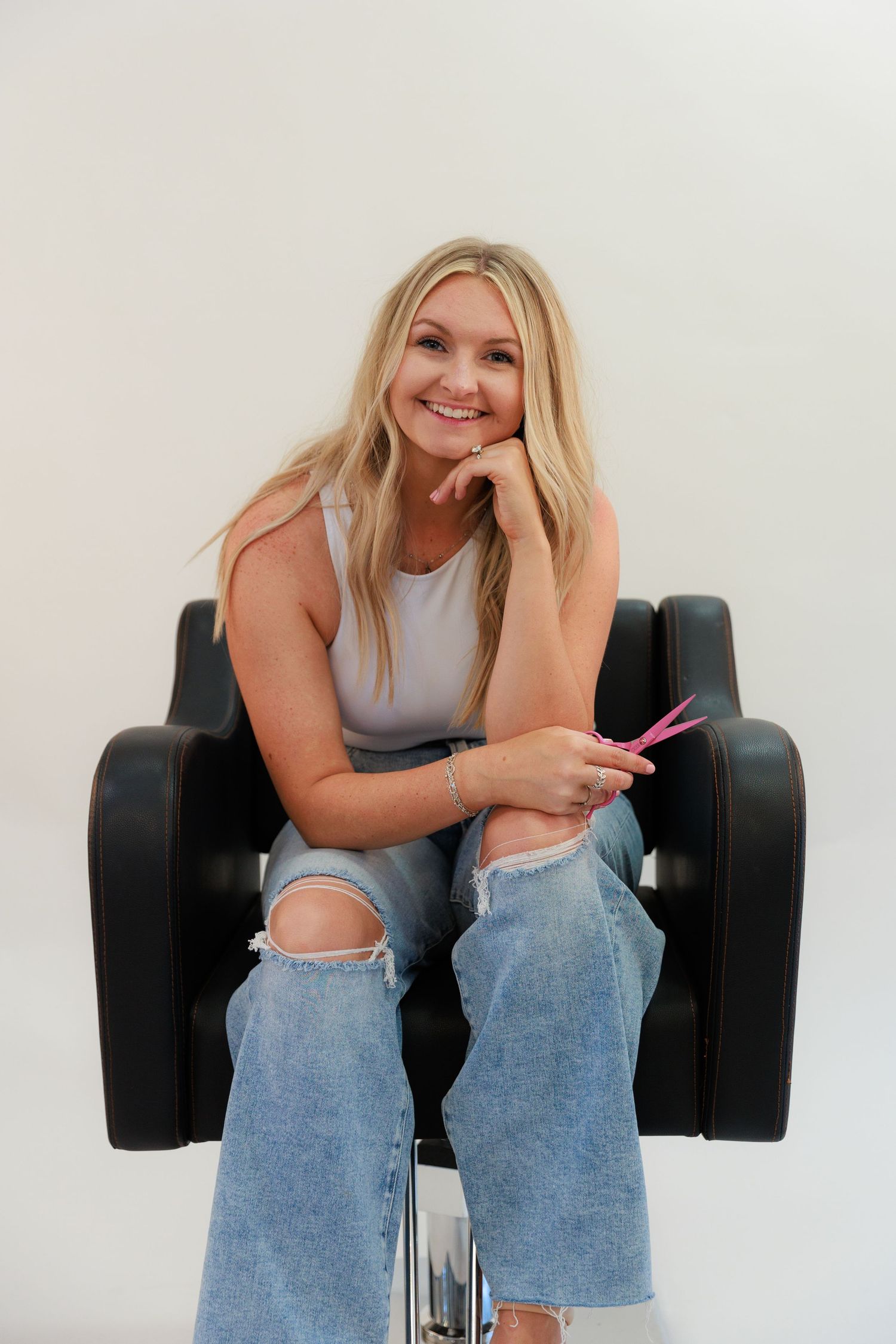 Woman in ripped jeans and tank top sits casually in black salon chair with pink tool in hand.