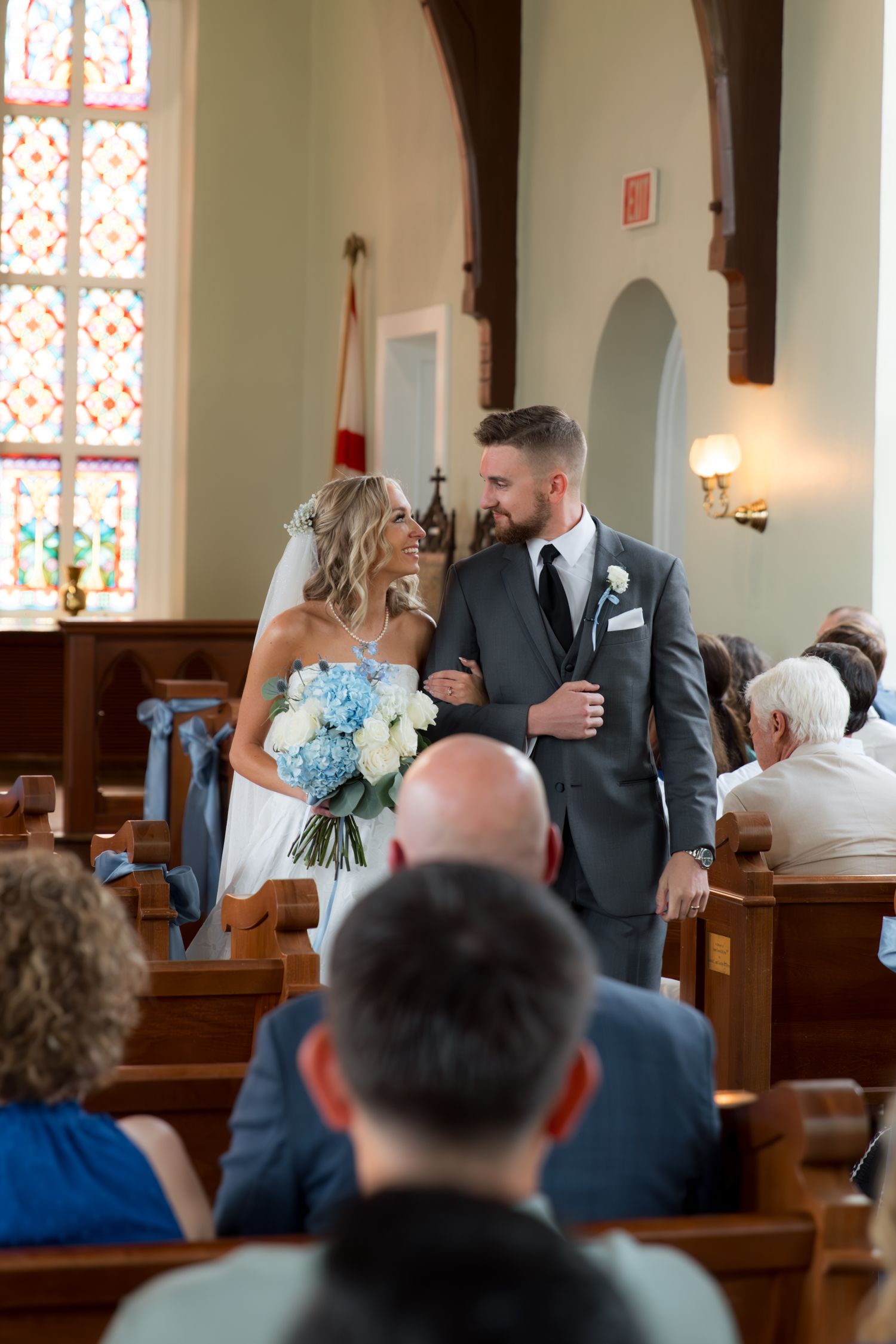 Wedding ceremony procession in a beautiful chapel with tall arched windows and decorative lighting.