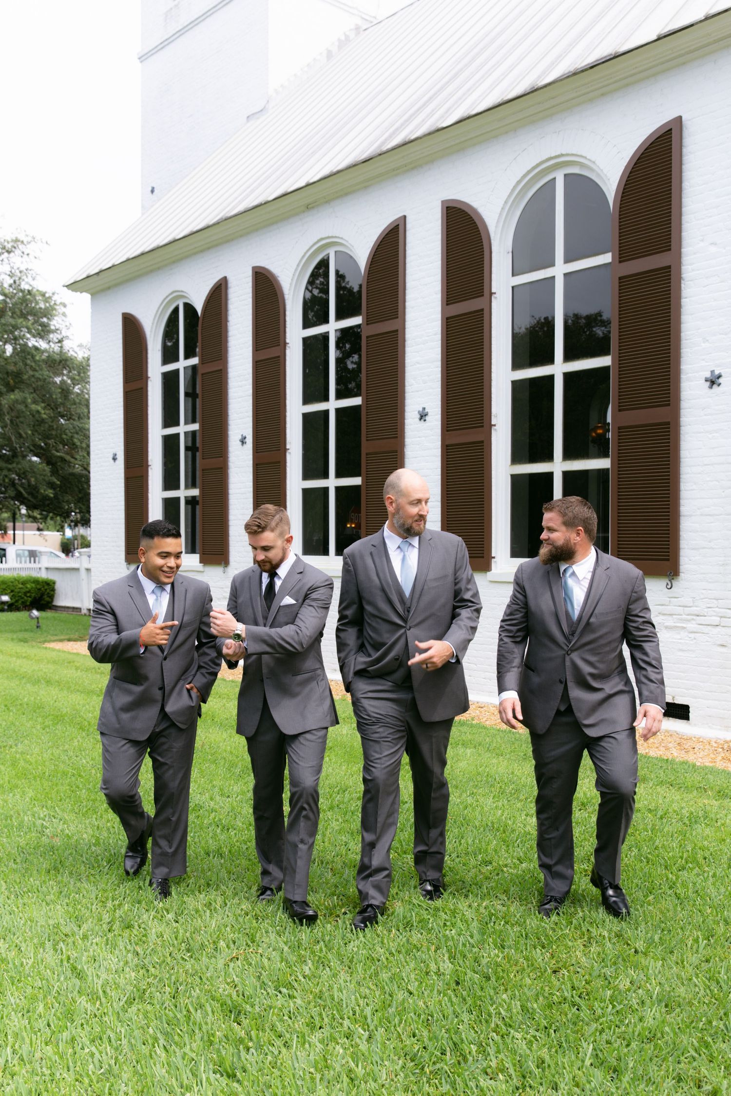 Groomsmen in matching gray suits walk and laugh together on church lawn.