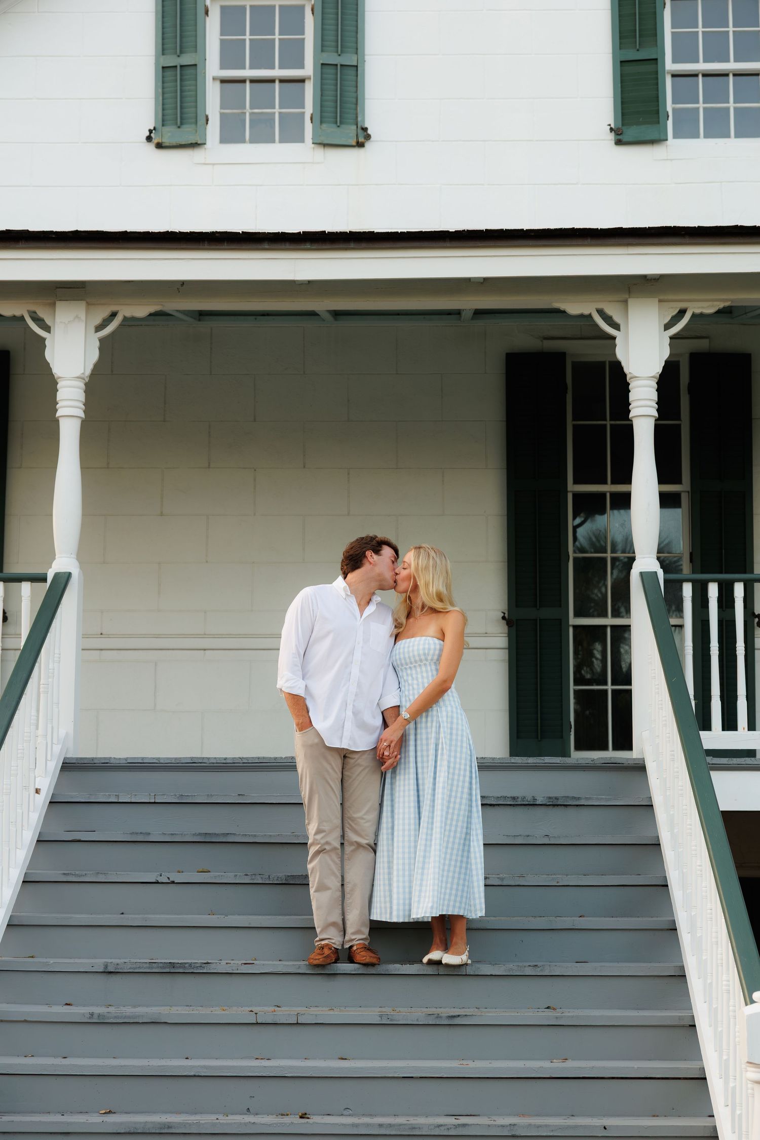 Couple sharing a kiss on gray porch steps of a white colonial home with green shutters.