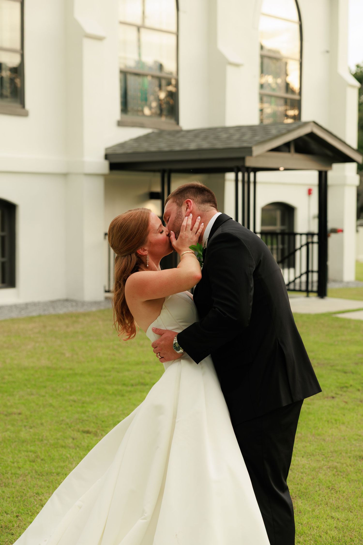 Bride and groom sharing romantic kiss on green lawn outside white wedding venue building.