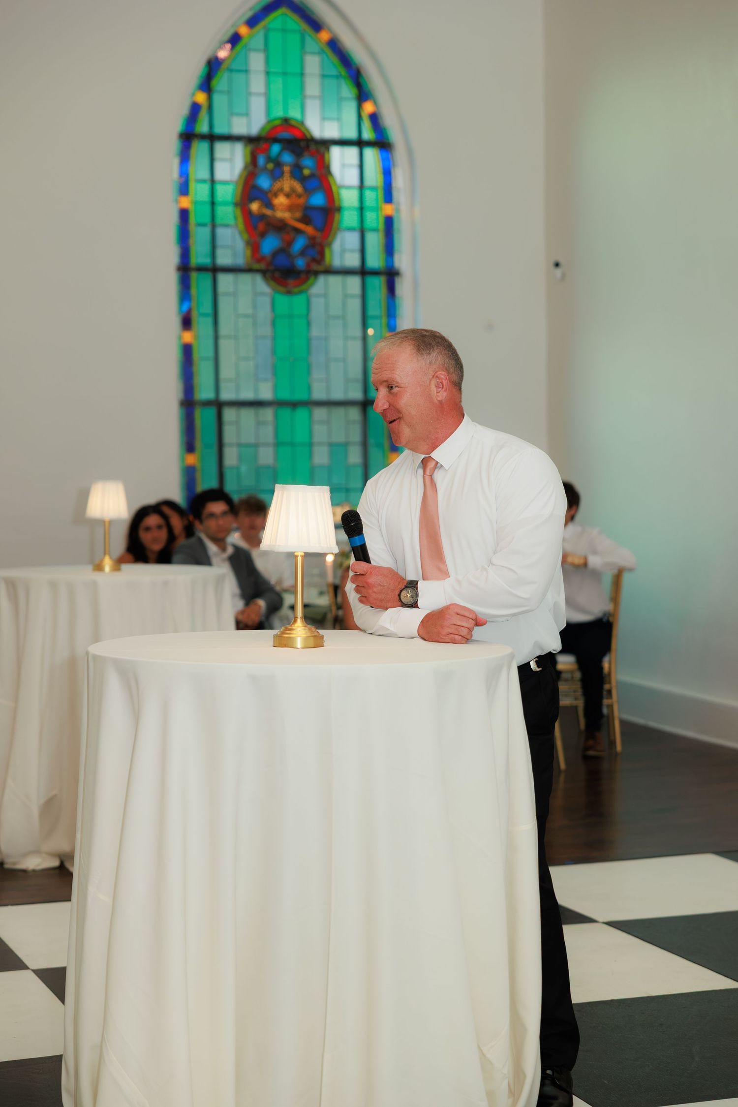 Man in white shirt giving speech at wedding reception with stained glass window backdrop.