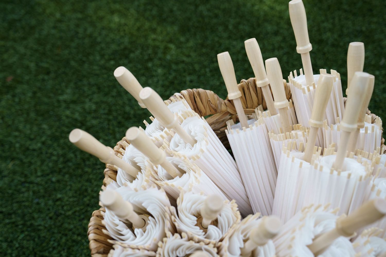 White parasols bundled in a wicker basket on green grass, perfect for outdoor wedding or garden party decor.