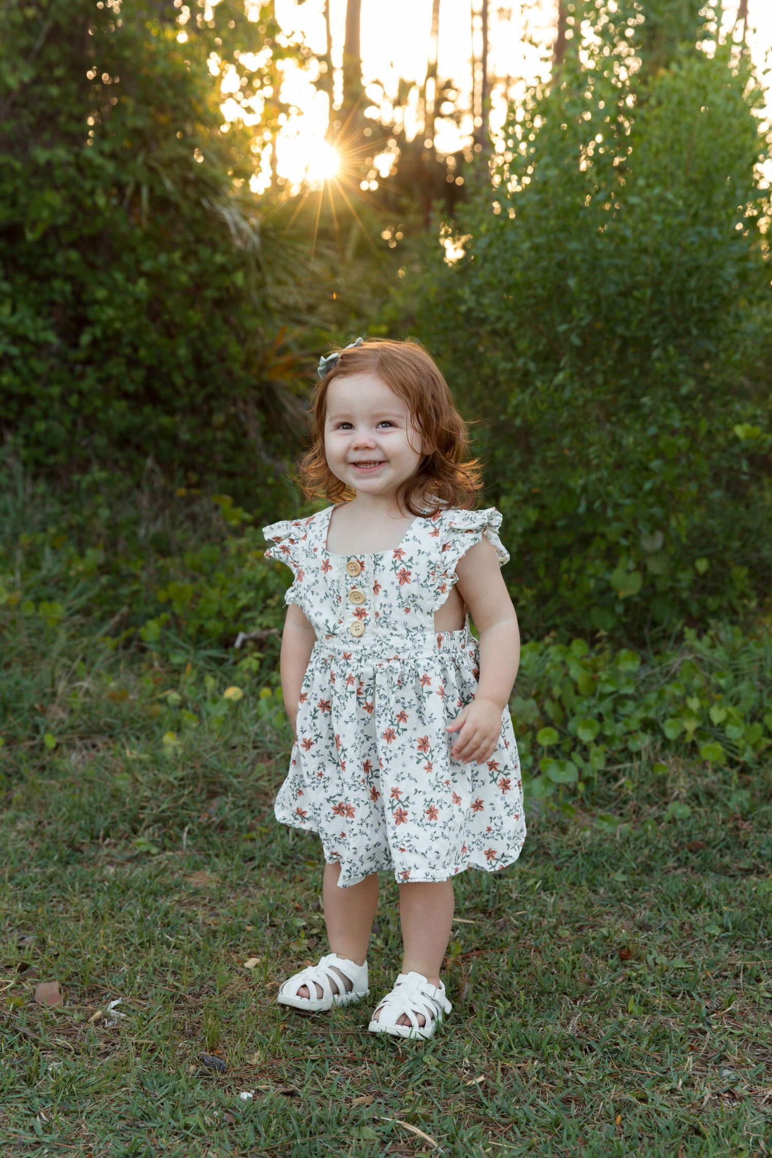A toddler in a floral dress and white sandals stands in a wooded area during golden hour sunset.