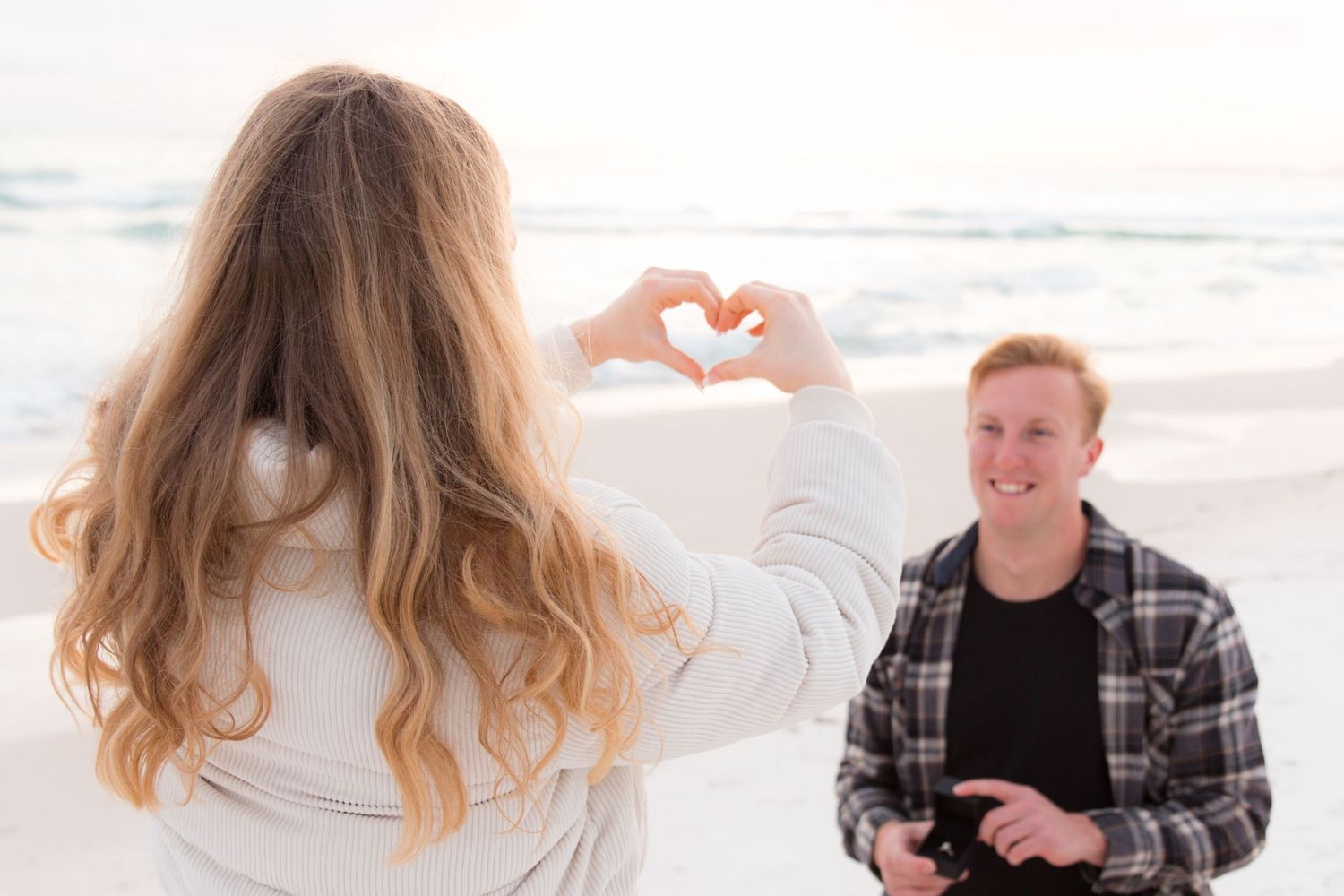 Woman with long blonde hair making heart shape with hands while standing on sandy beach at sunset.
