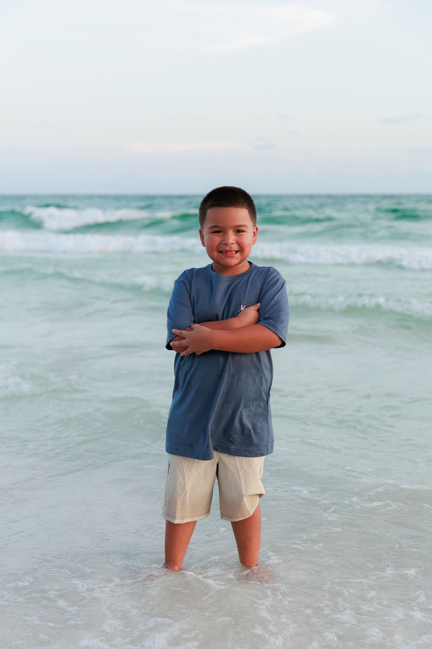 Young boy in blue shirt and khaki shorts stands confidently in shallow ocean water with waves behind him.
