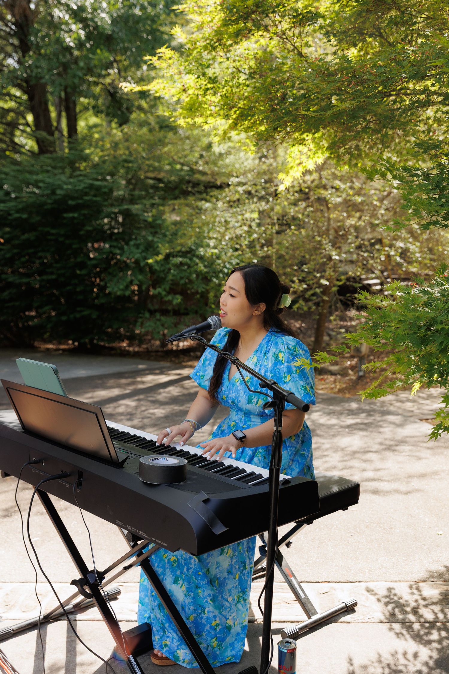 Young Asian woman in blue floral dress performing on keyboard at outdoor garden event.