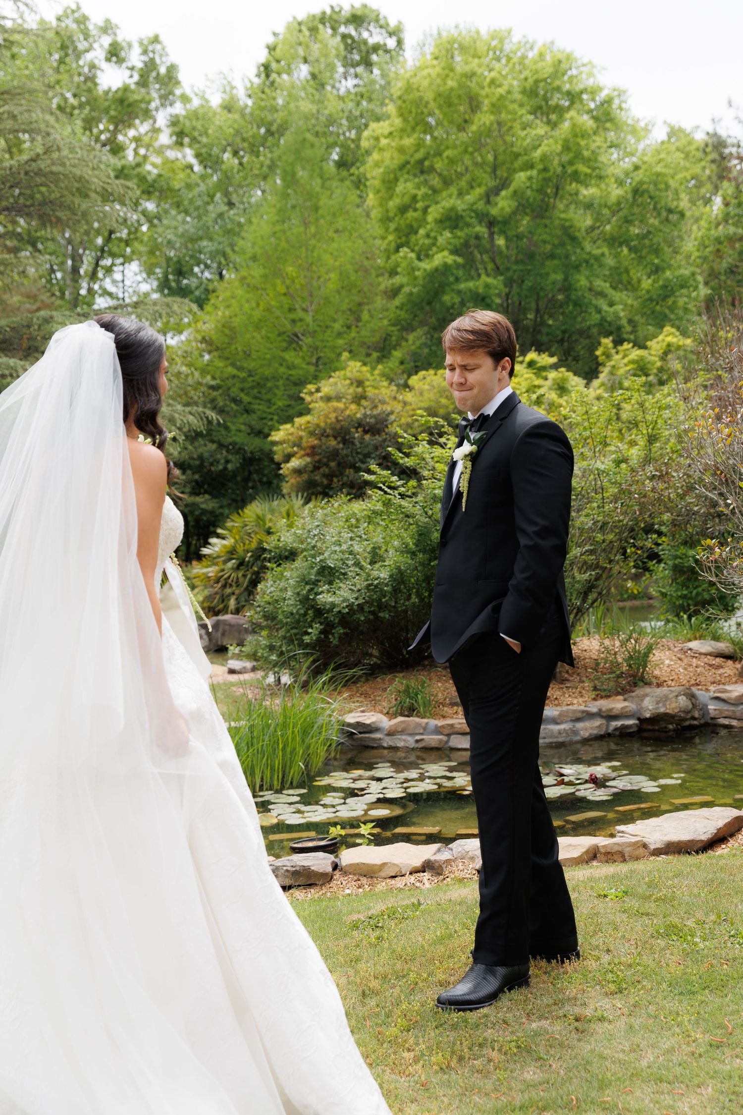 Groom in black tuxedo smiles at bride in white wedding gown during first look at garden venue.