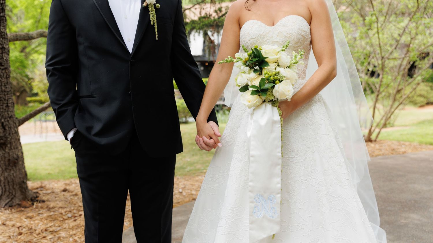 Bride and groom holding hands outdoors, bride wearing strapless white gown with veil holding white floral bouquet.