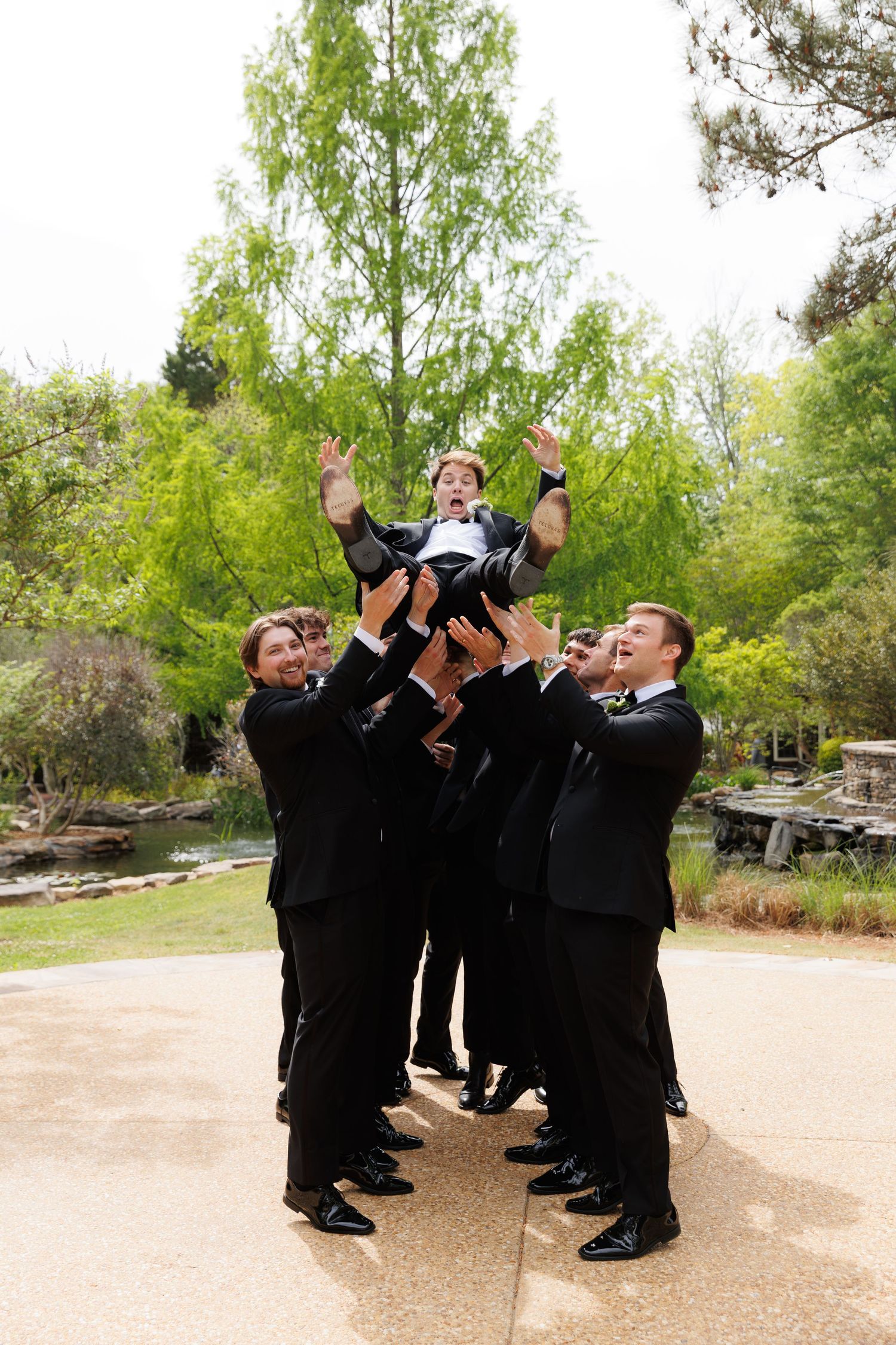 Groomsmen in black tuxedos tossing groom into the air outdoors at a wedding.