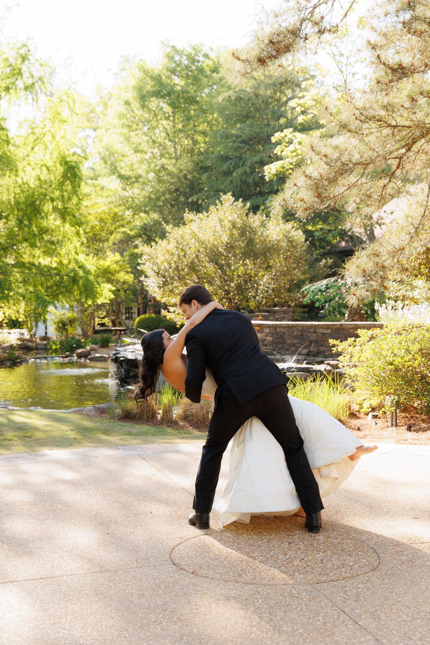 Groom dips bride in white wedding gown during romantic outdoor dance near a garden pond.