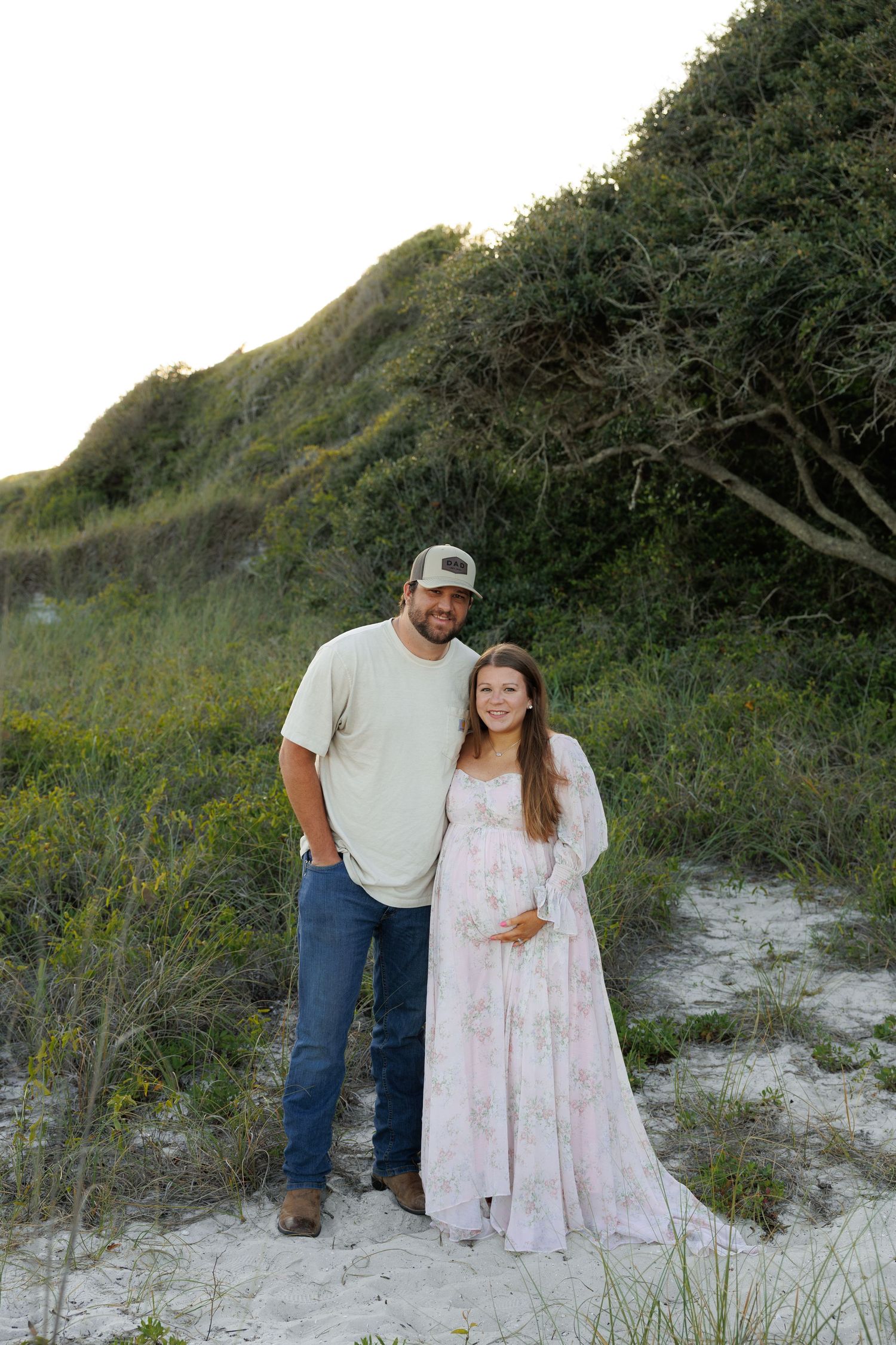 Couple poses on sandy beach dunes, pregnant woman in floral maxi dress with partner in cap and jeans.