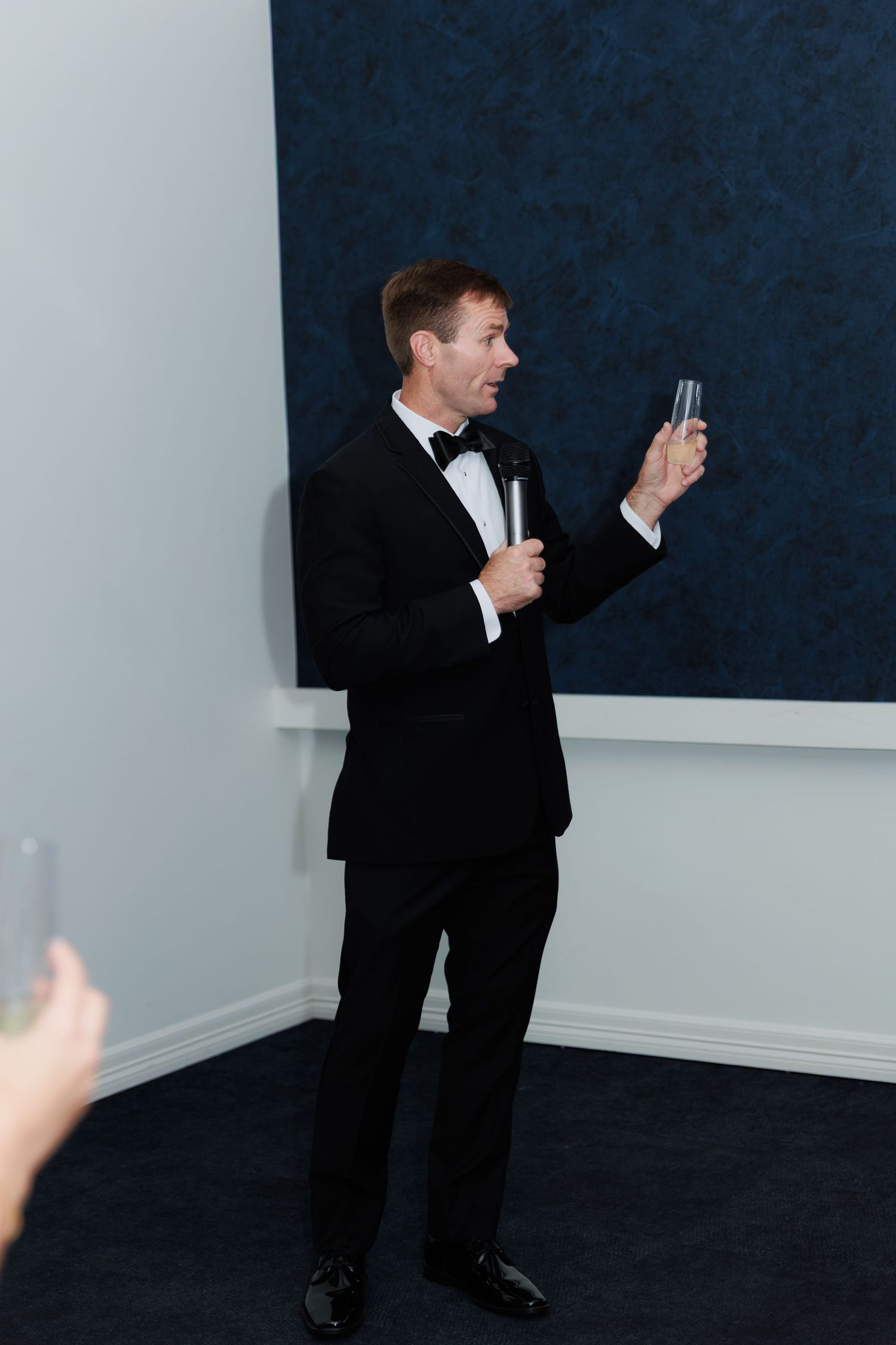 A person in a formal black tuxedo and bowtie stands holding a microphone and phone during an event.