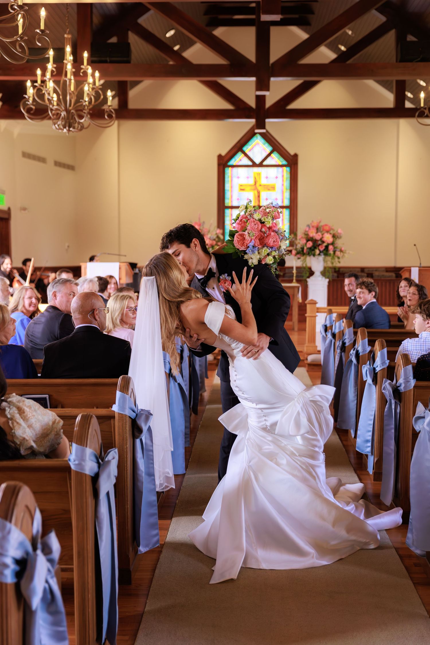Newlyweds share their first kiss in a chapel with exposed wooden beams and stained glass windows.