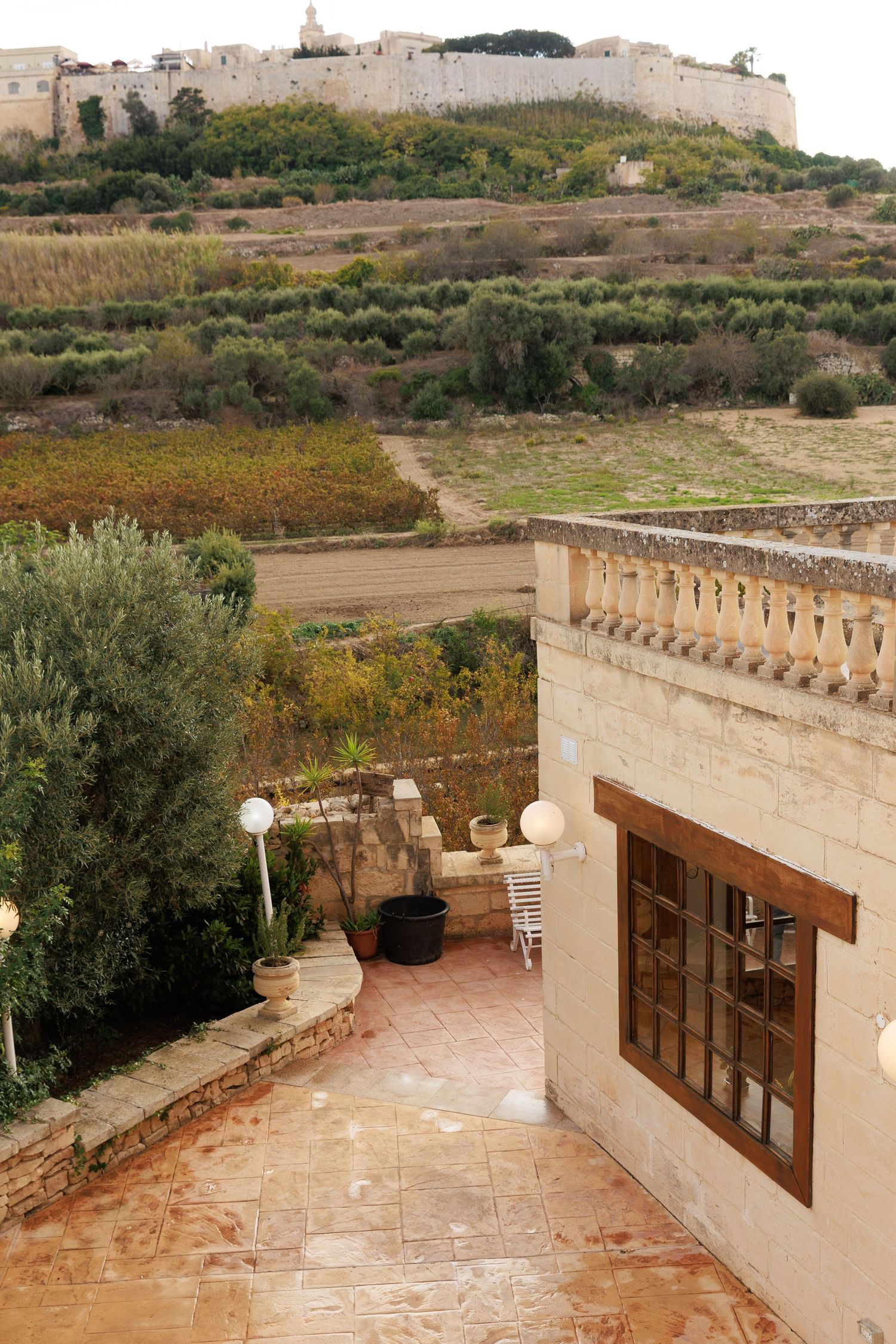 Mediterranean-style terrace with stone walls and wooden windows overlooking a rural landscape with olive trees.