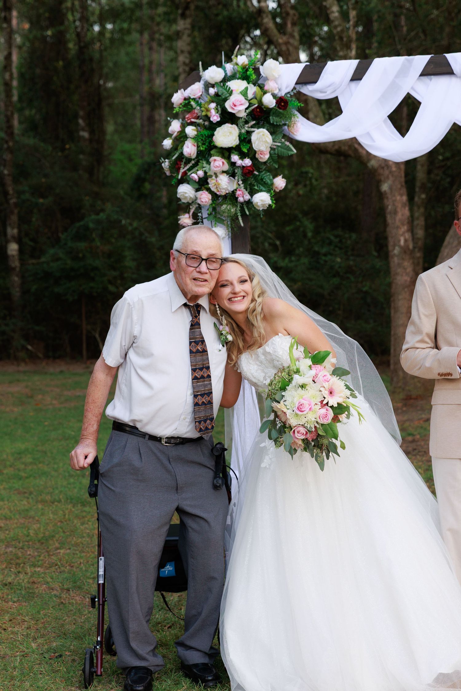 A wedding scene in a wooded outdoor setting with floral decorations and people in formal attire.