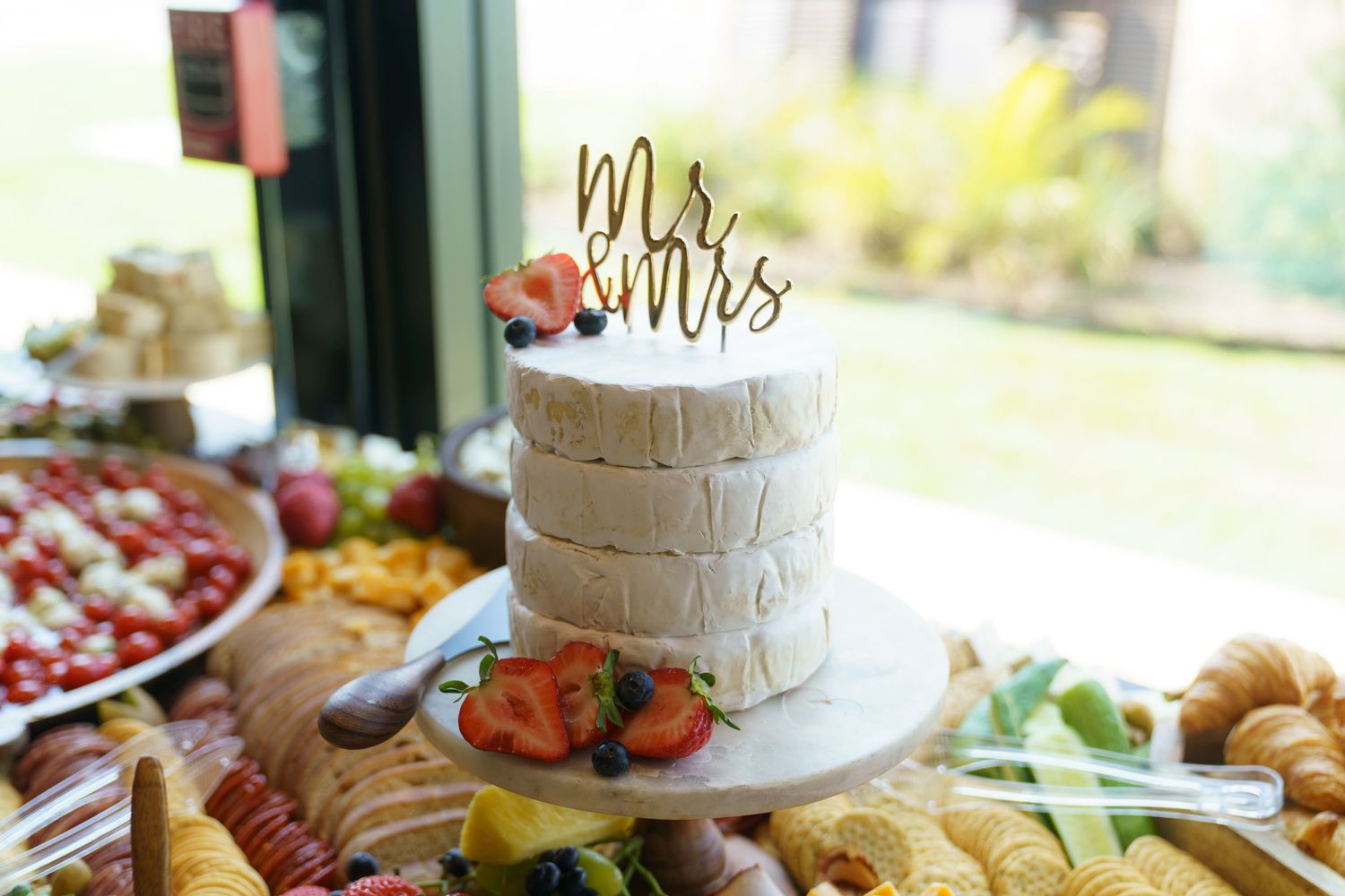 A wedding reception table displays an elegant spread of appetizers, cheese boards, and a white tiered cake.