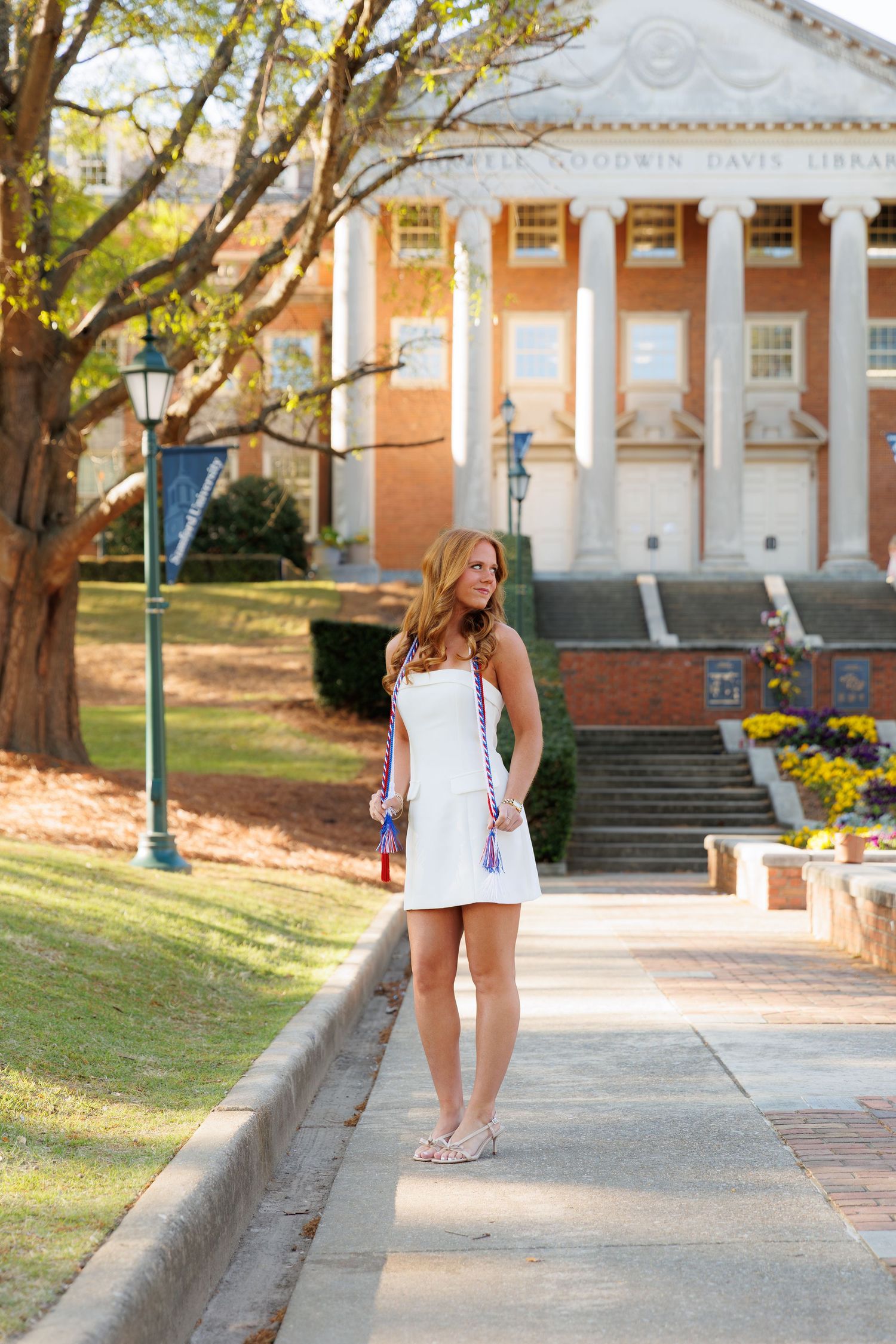 Young woman in white dress with colorful sashes stands on campus walkway in front of a brick library building.