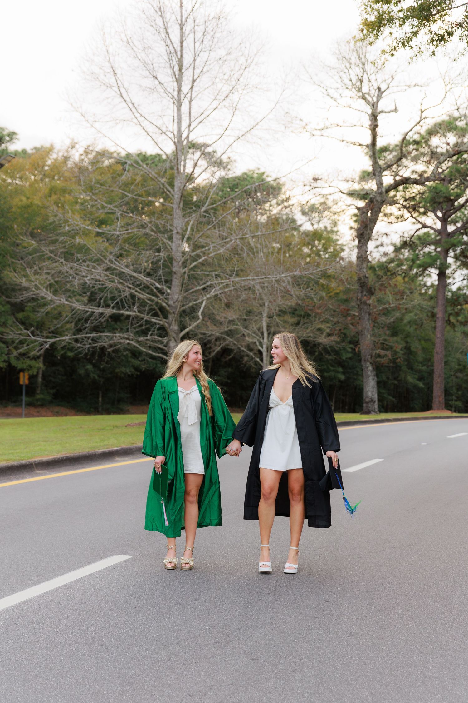 Two female graduates in green and black robes hold hands walking down a tree-lined road during graduation photo session.