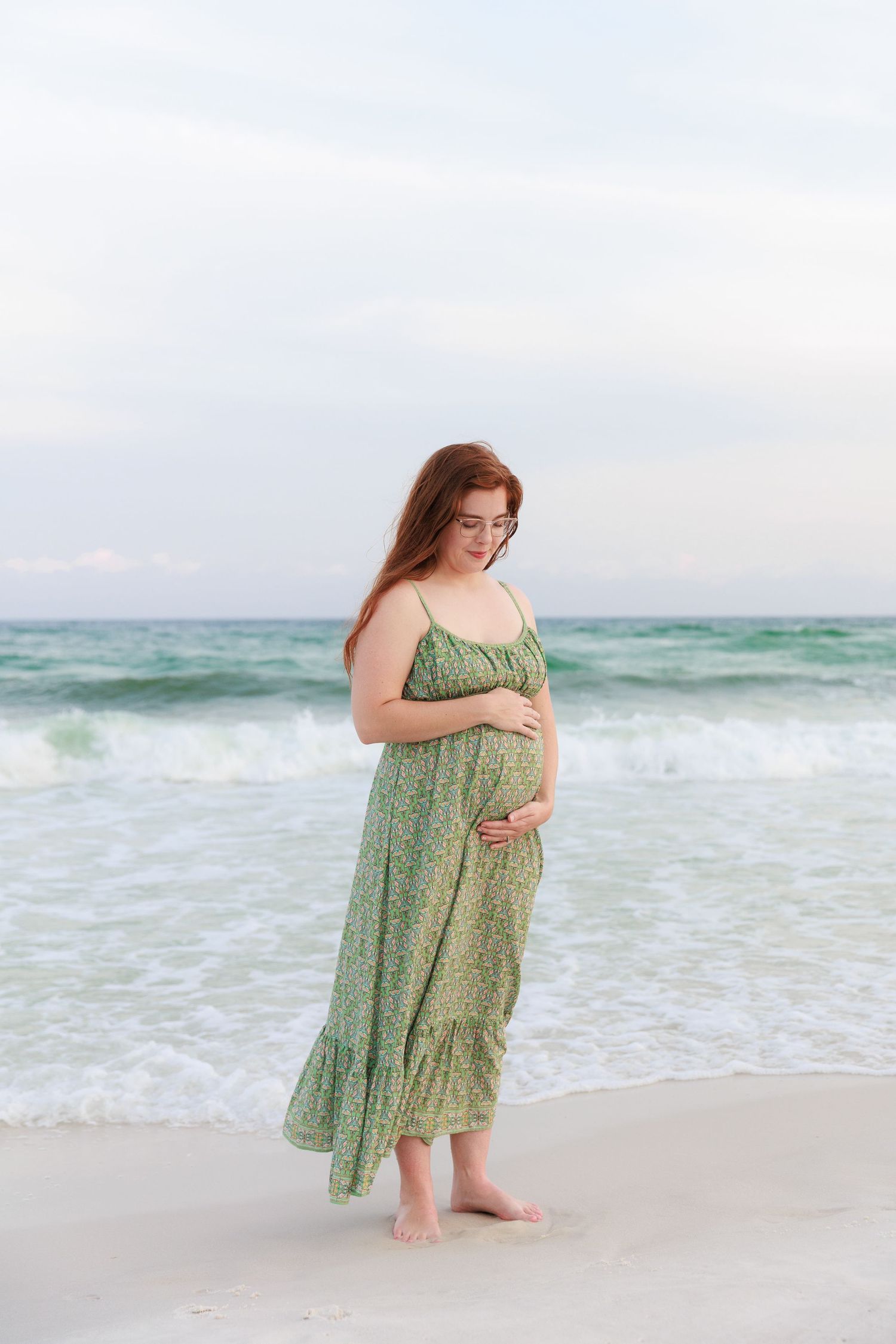 A woman in a flowy green dress walks along a serene beach with turquoise waves in the background.
