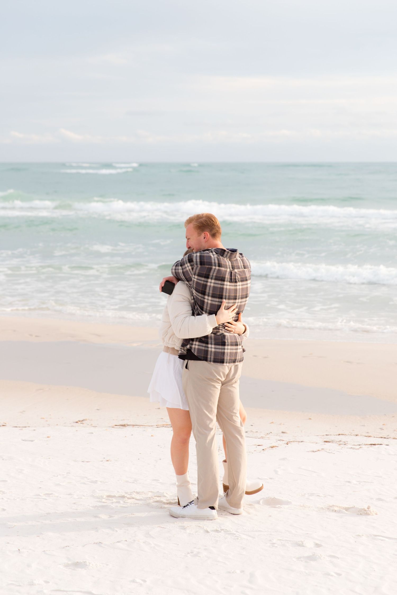 A couple shares an intimate moment on a sandy beach with gentle waves and cloudy skies in the distance.