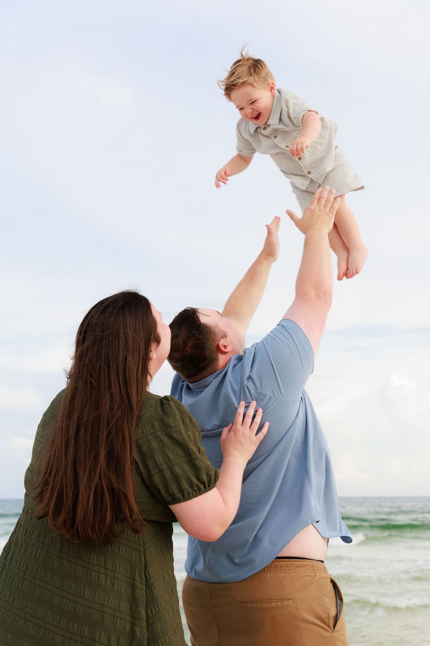 A couple stands by the ocean, lifting and playing with a baby in a sequence of joyful moments at the beach.