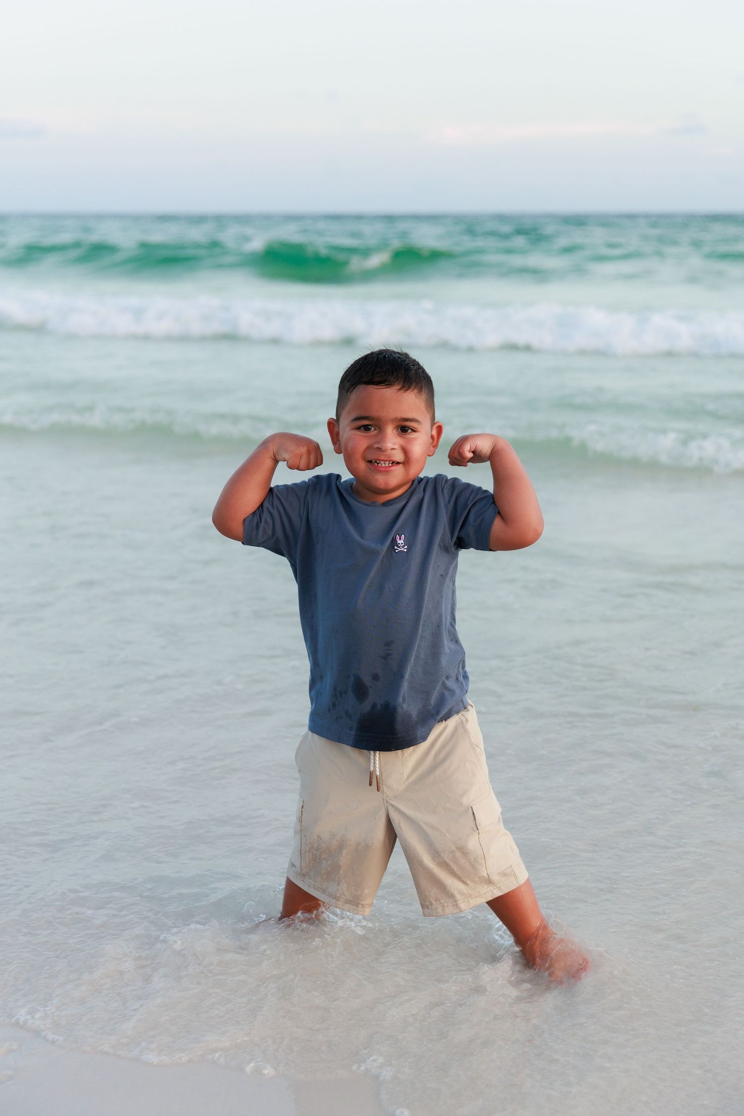 Child poses energetically at the beach with waves and turquoise ocean water in the background.