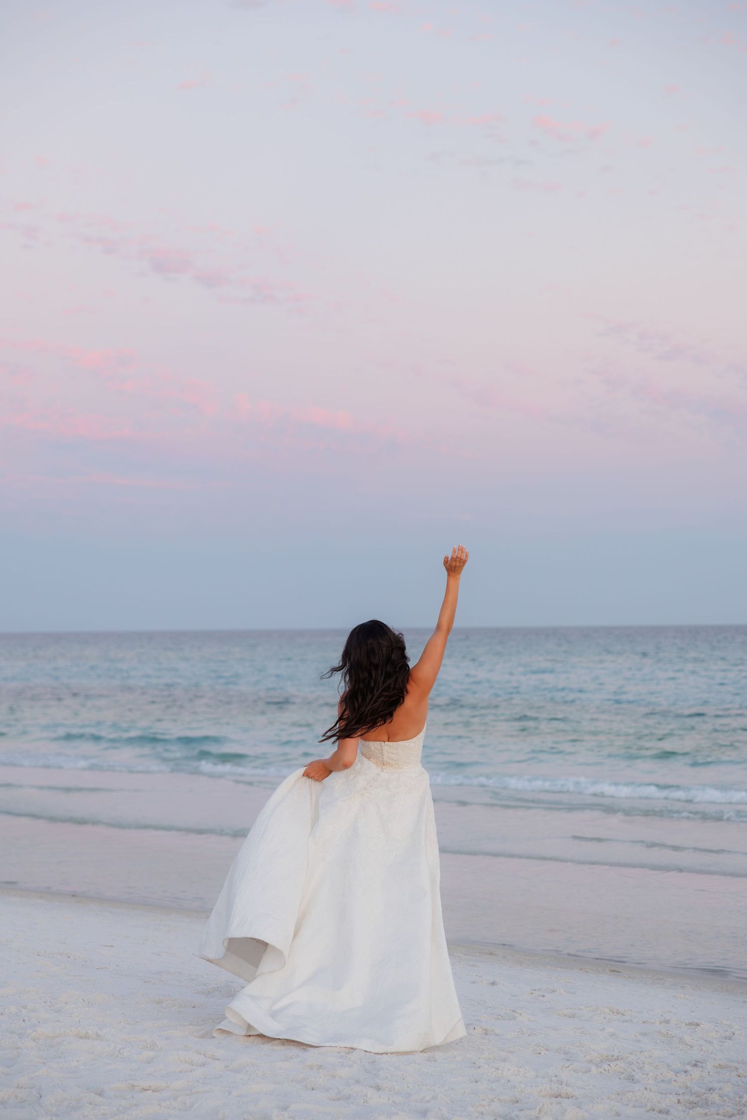 Bride in strapless white gown raises fist triumphantly on beach at sunset with pastel pink and blue sky.