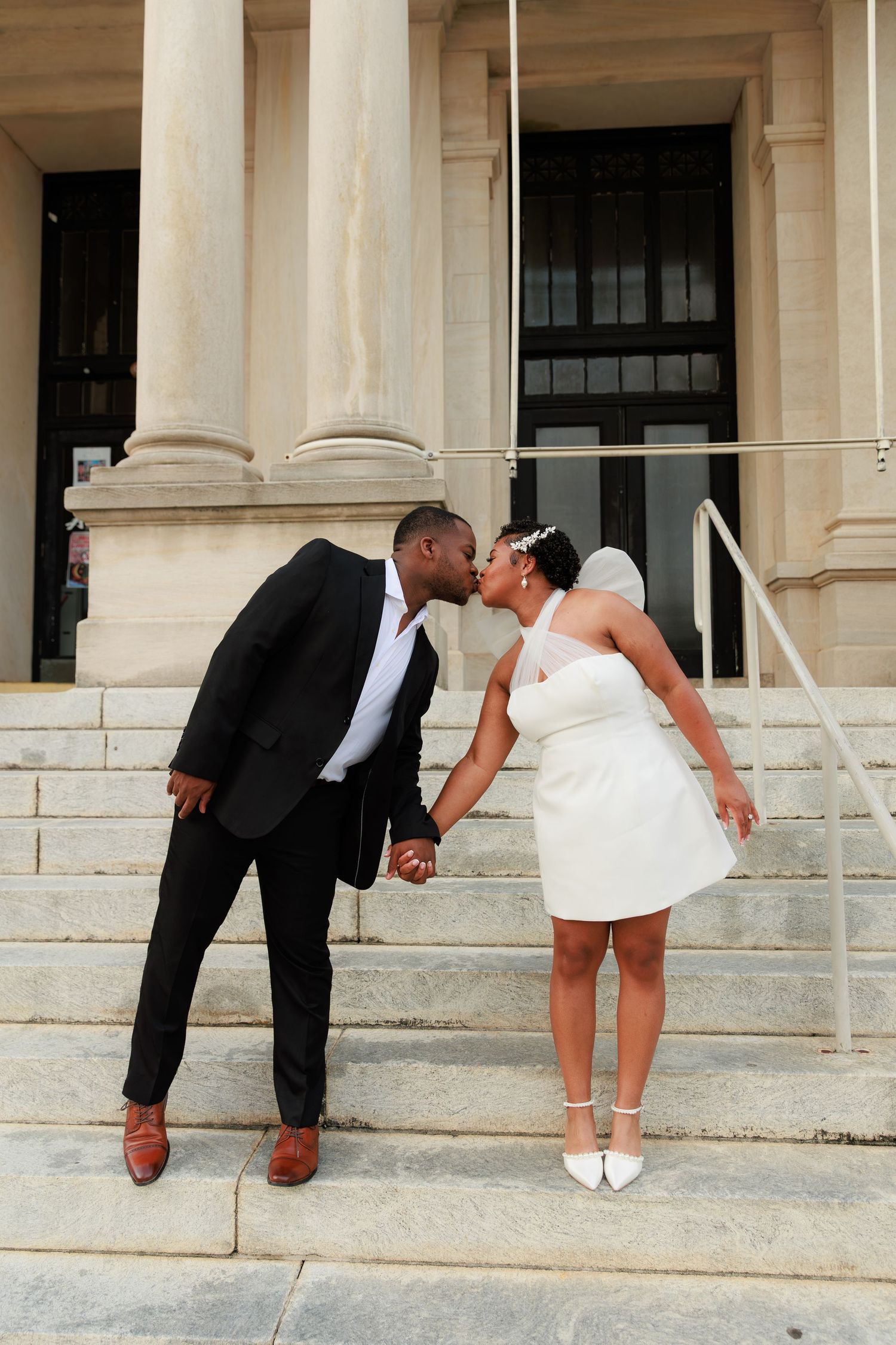 Newlywed couple kissing on courthouse steps, groom in black suit, bride in white mini dress and heels.