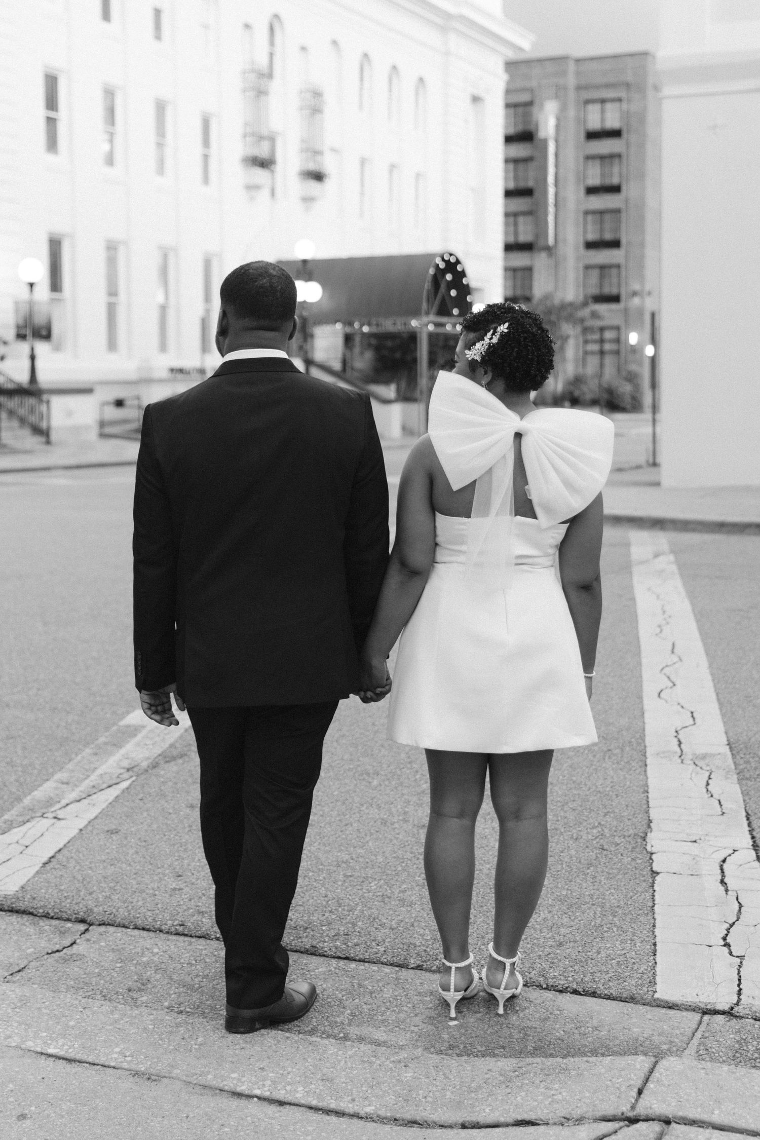 Black and white photo of a bride in a short white dress with bow and groom in suit walking hand in hand outdoors.
