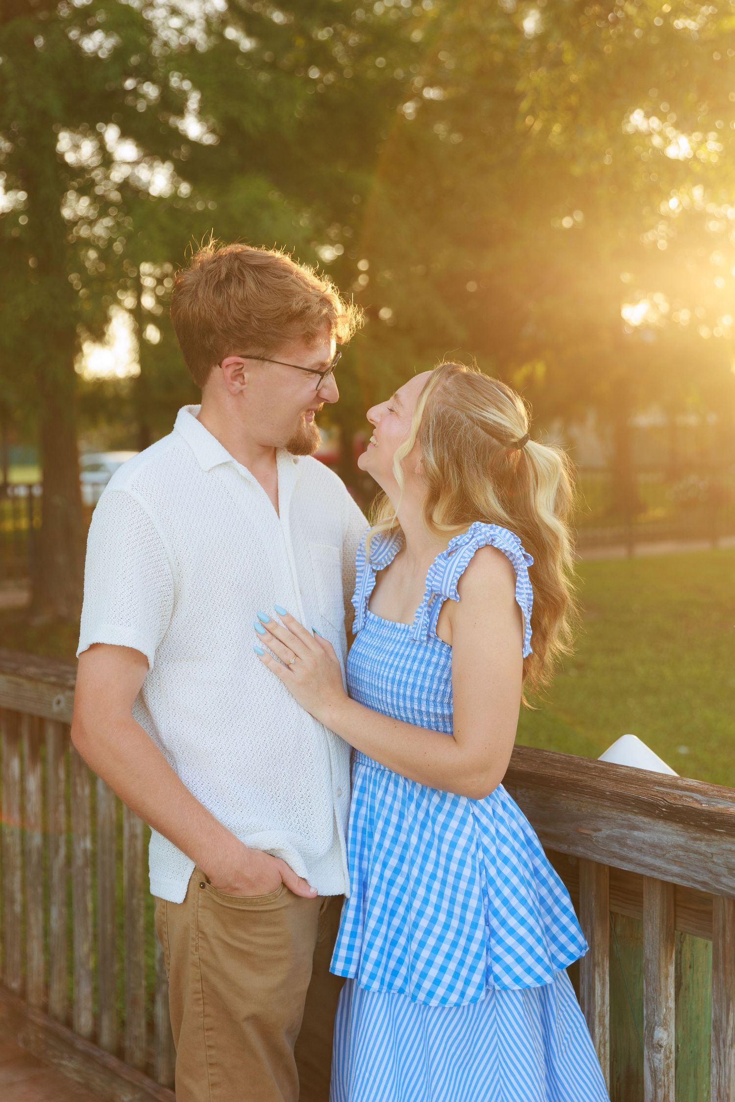A happy couple gazes at each other on a wooden bridge, bathed in warm golden sunset light, she wears a blue gingham dress.